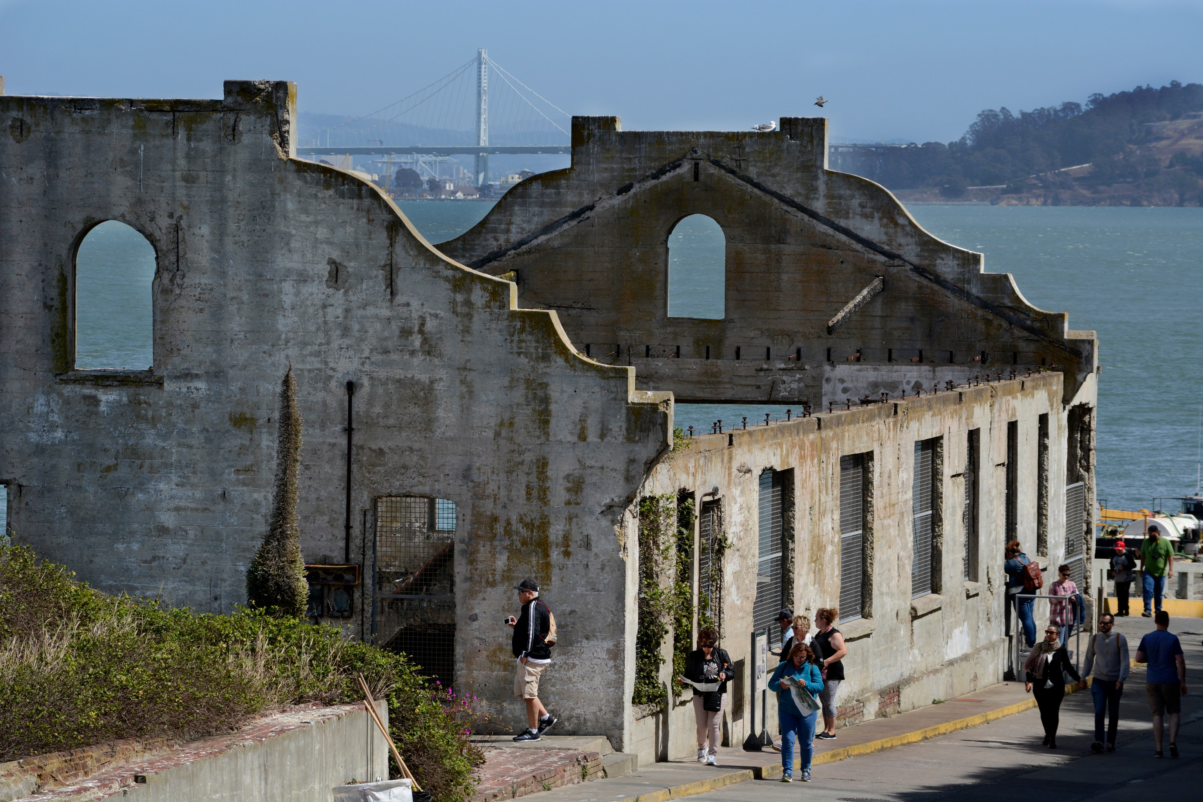 A weathered concrete building stands by the water, with people walking around it. In the background, a bridge spans across the bay.