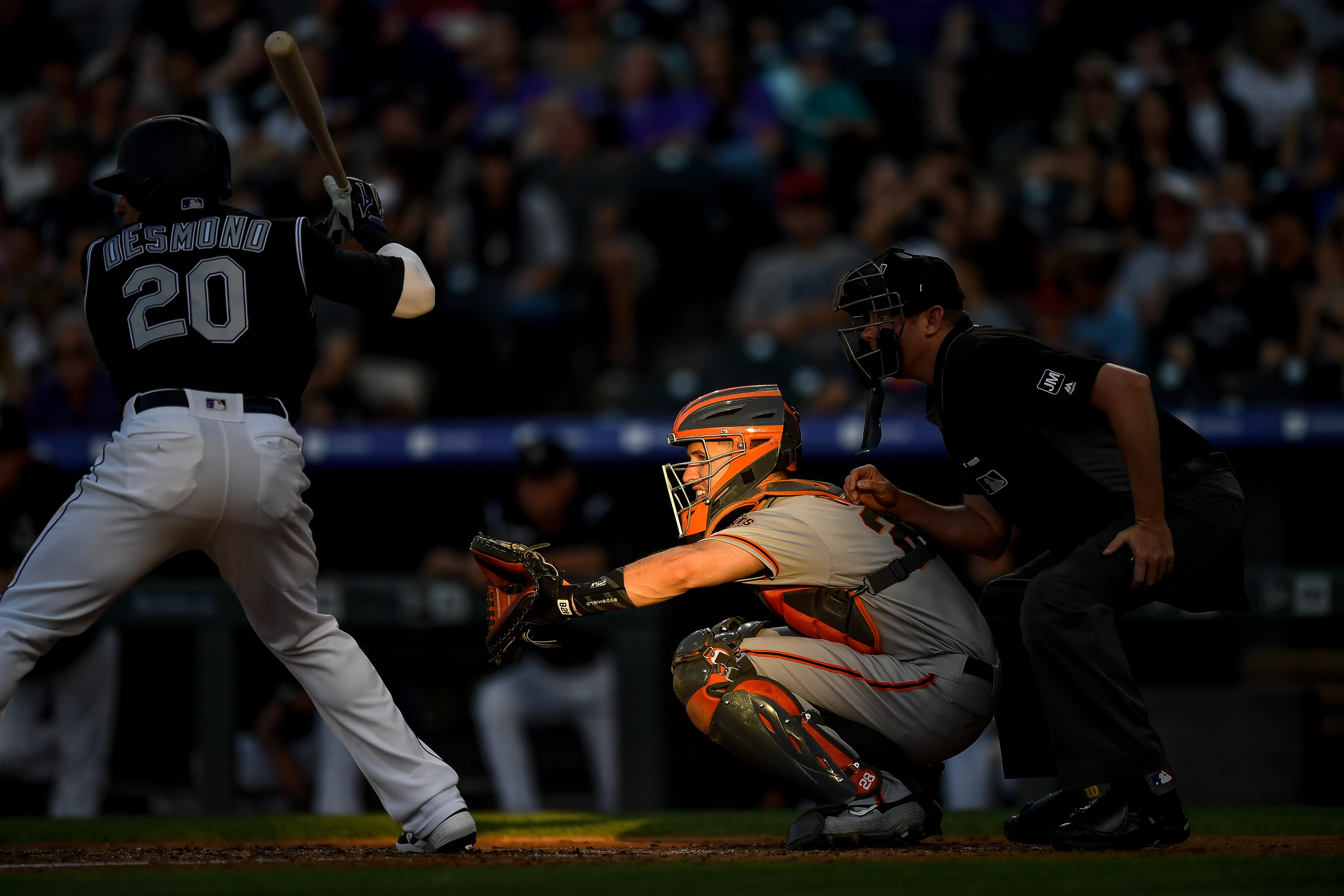 A baseball batter prepares to swing as the catcher crouches behind home plate, with an umpire leaning in. The scene is partially shadowed, emphasizing the players.