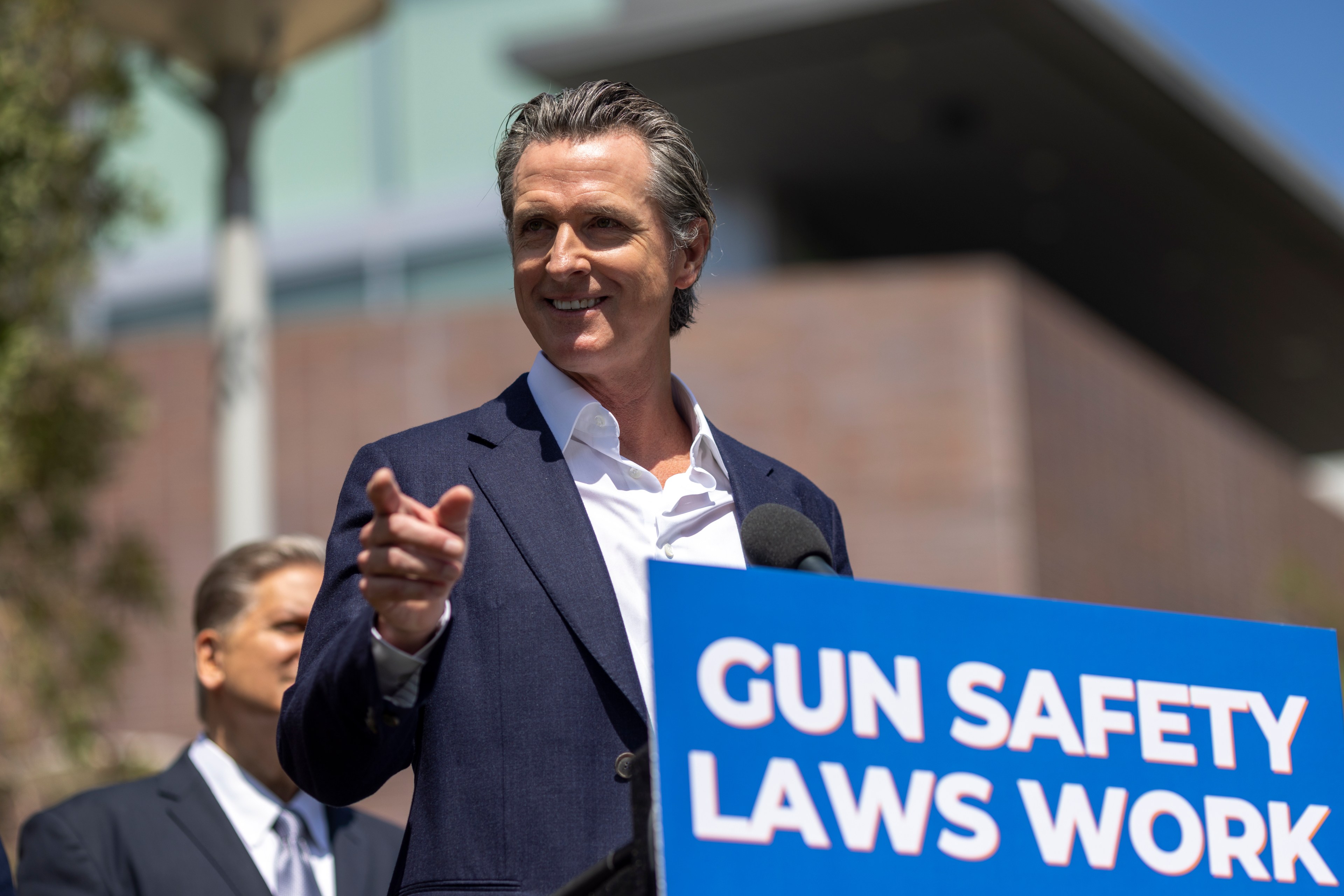 A man in a suit speaks at a podium with a sign reading "Gun Safety Laws Work." Another person stands in the background, and a building is visible behind them.