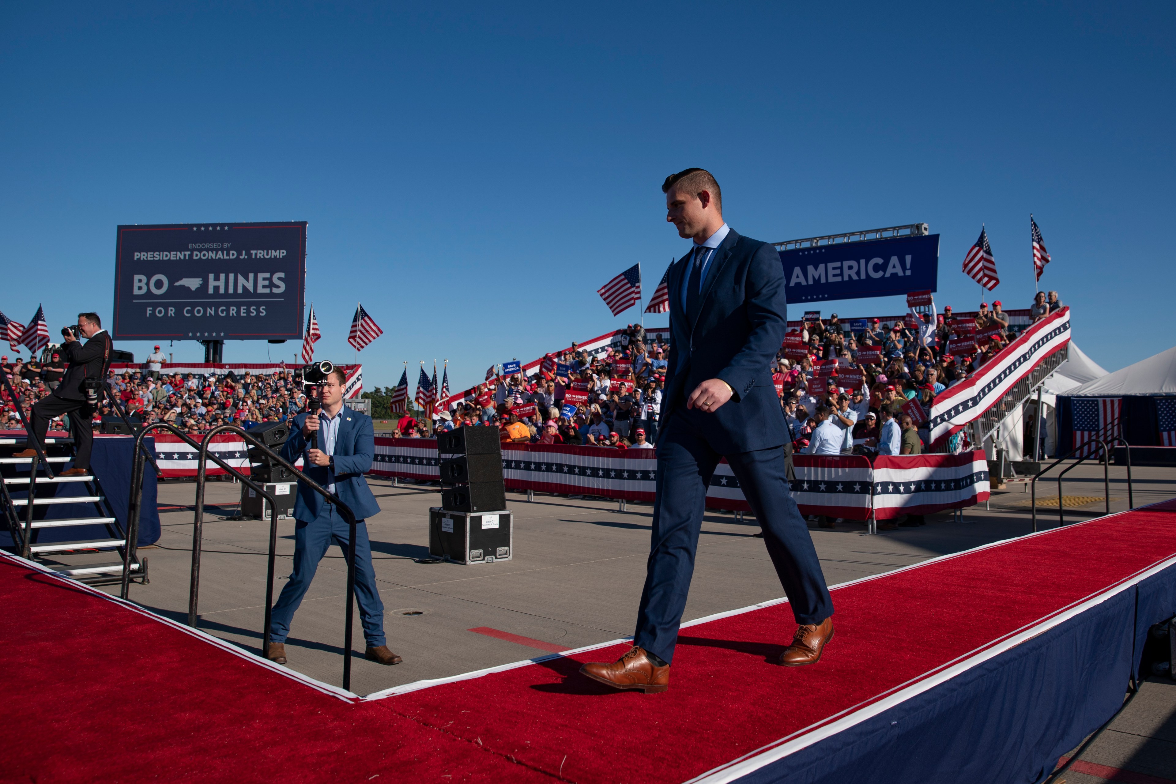 A man in a blue suit walks on a red carpeted stage at a rally. The crowd, holding American flags, is seated in bleachers under a clear blue sky.