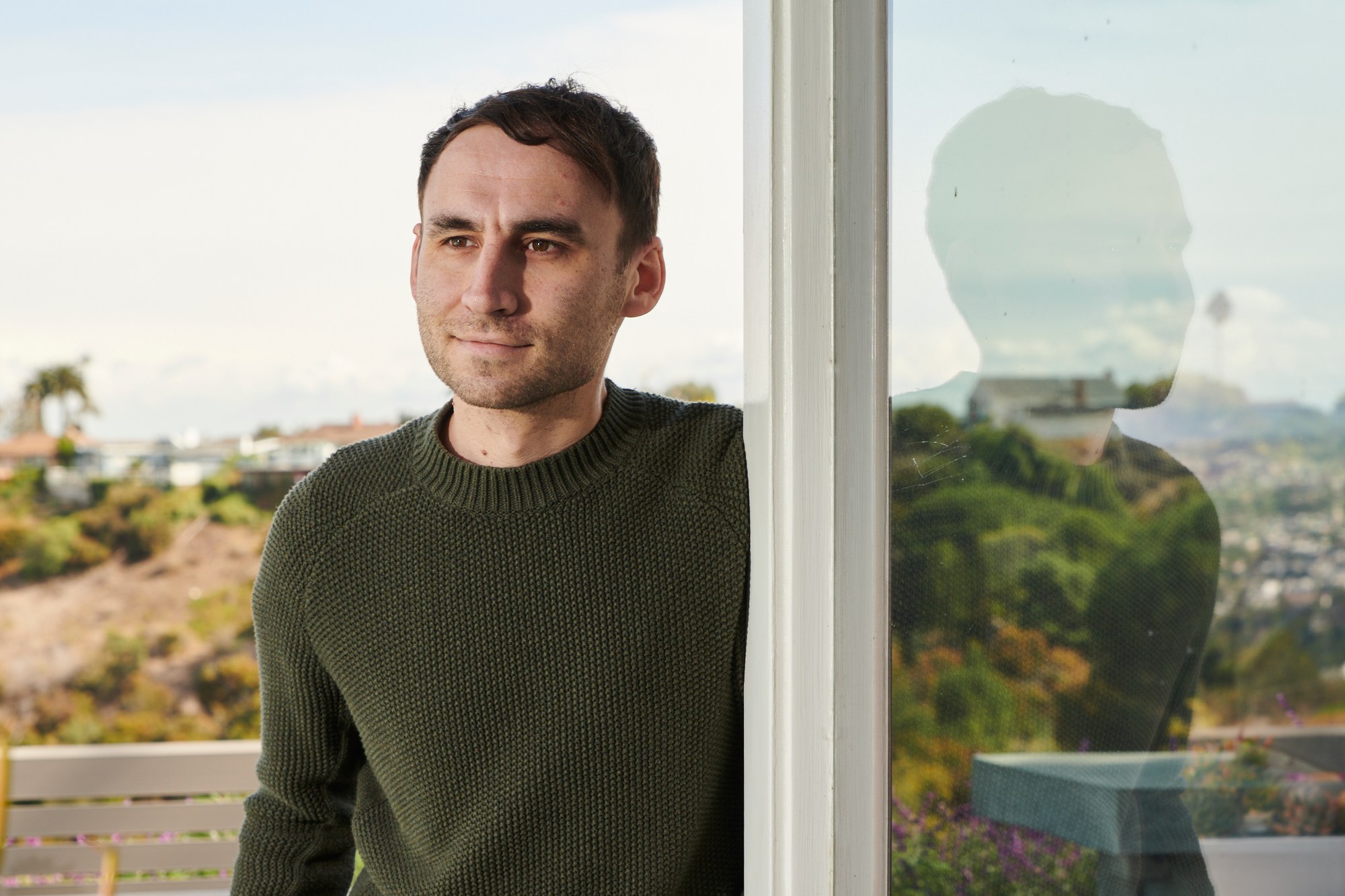 A person in a green sweater stands by a glass wall, hands in pockets. There's a reflection in the glass and a blurred outdoor view with plants and a clear sky.