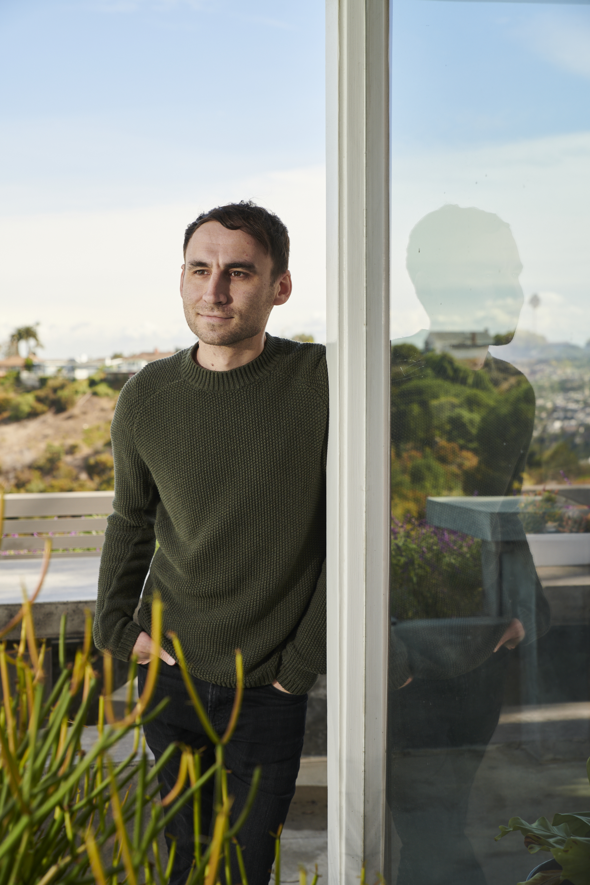 A person in a green sweater stands by a glass wall, hands in pockets. There's a reflection in the glass and a blurred outdoor view with plants and a clear sky.
