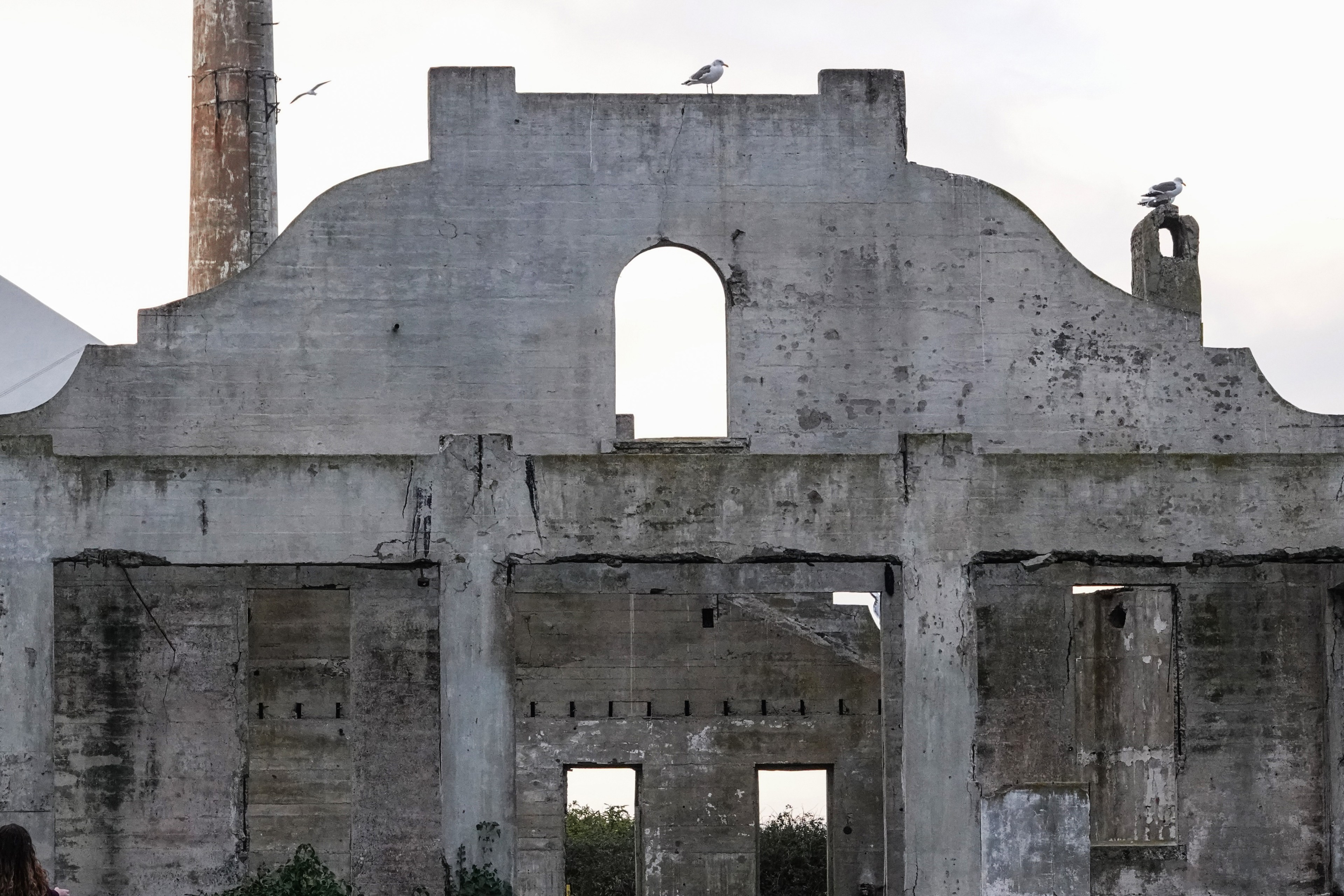 A weathered concrete structure with an arched opening at the top, accompanied by a few seagulls perched on it, and a tall chimney in the background.