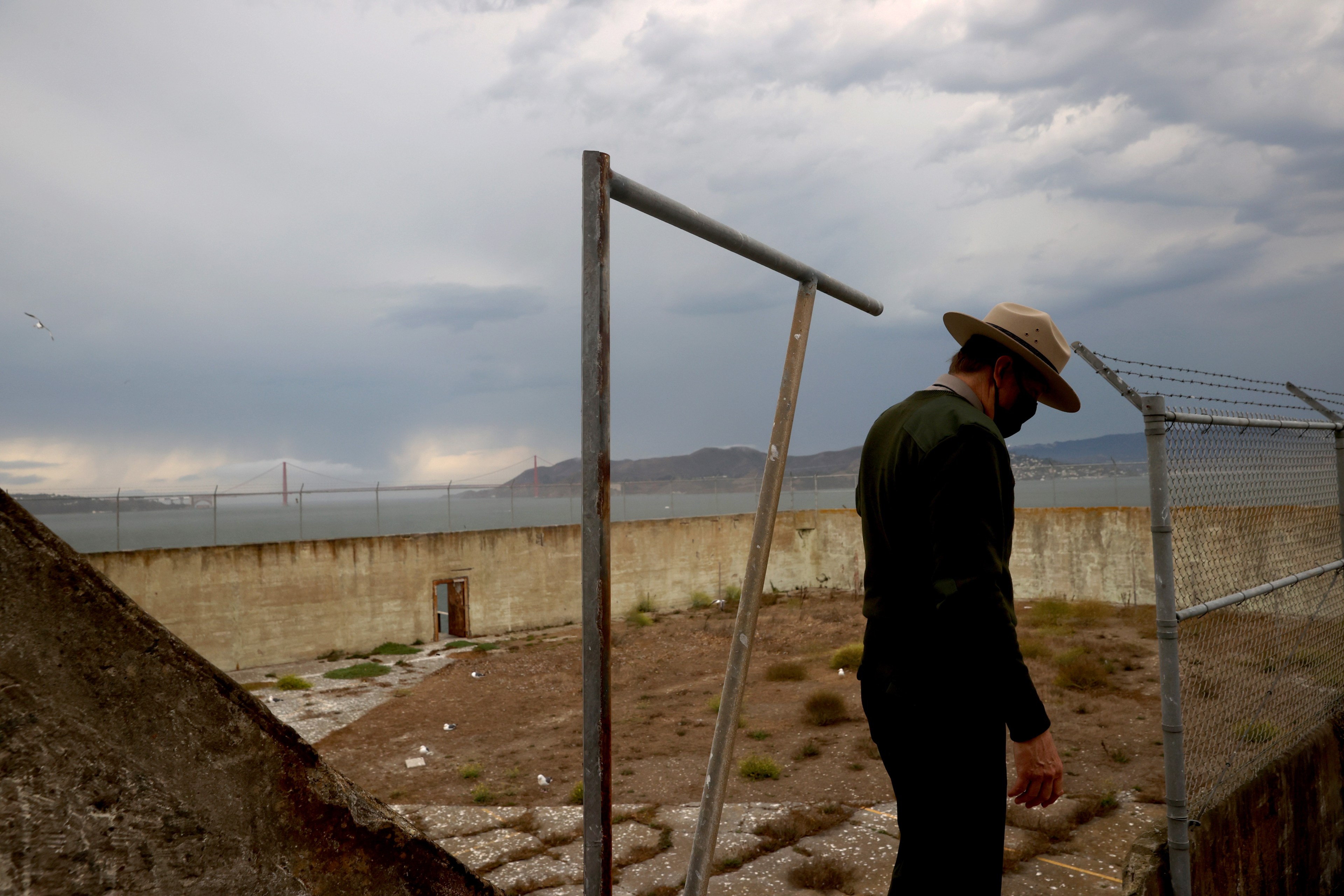 A person in a wide-brimmed hat and mask stands beside a fenced area, overlooking a body of water and distant bridge under cloudy skies.