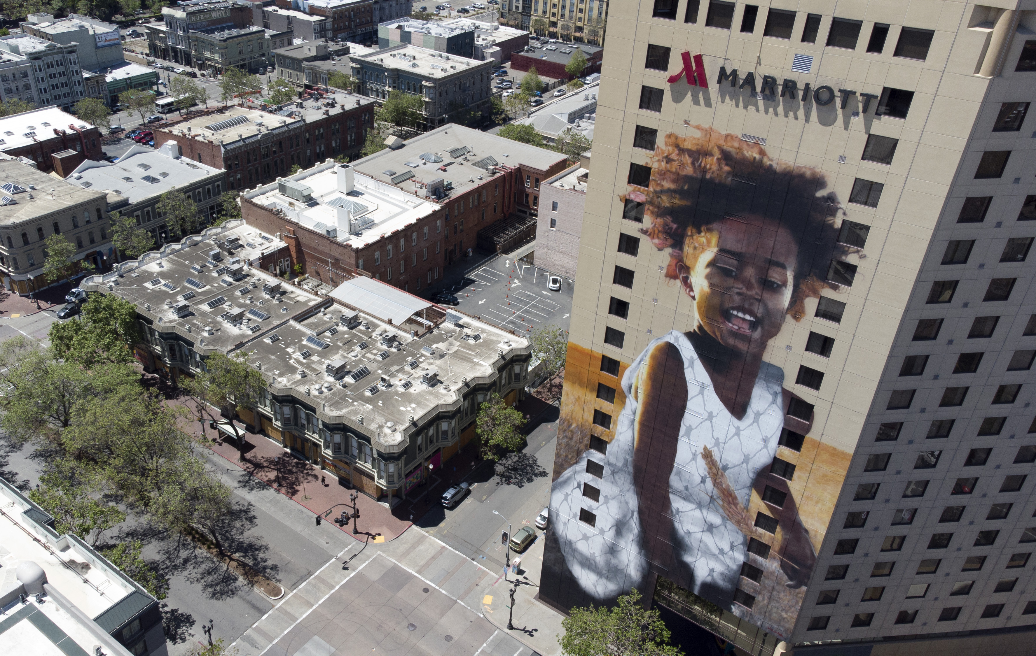 The image shows a cityscape with a tall hotel building featuring a large mural of a smiling child in a white dress, next to historic-style buildings.
