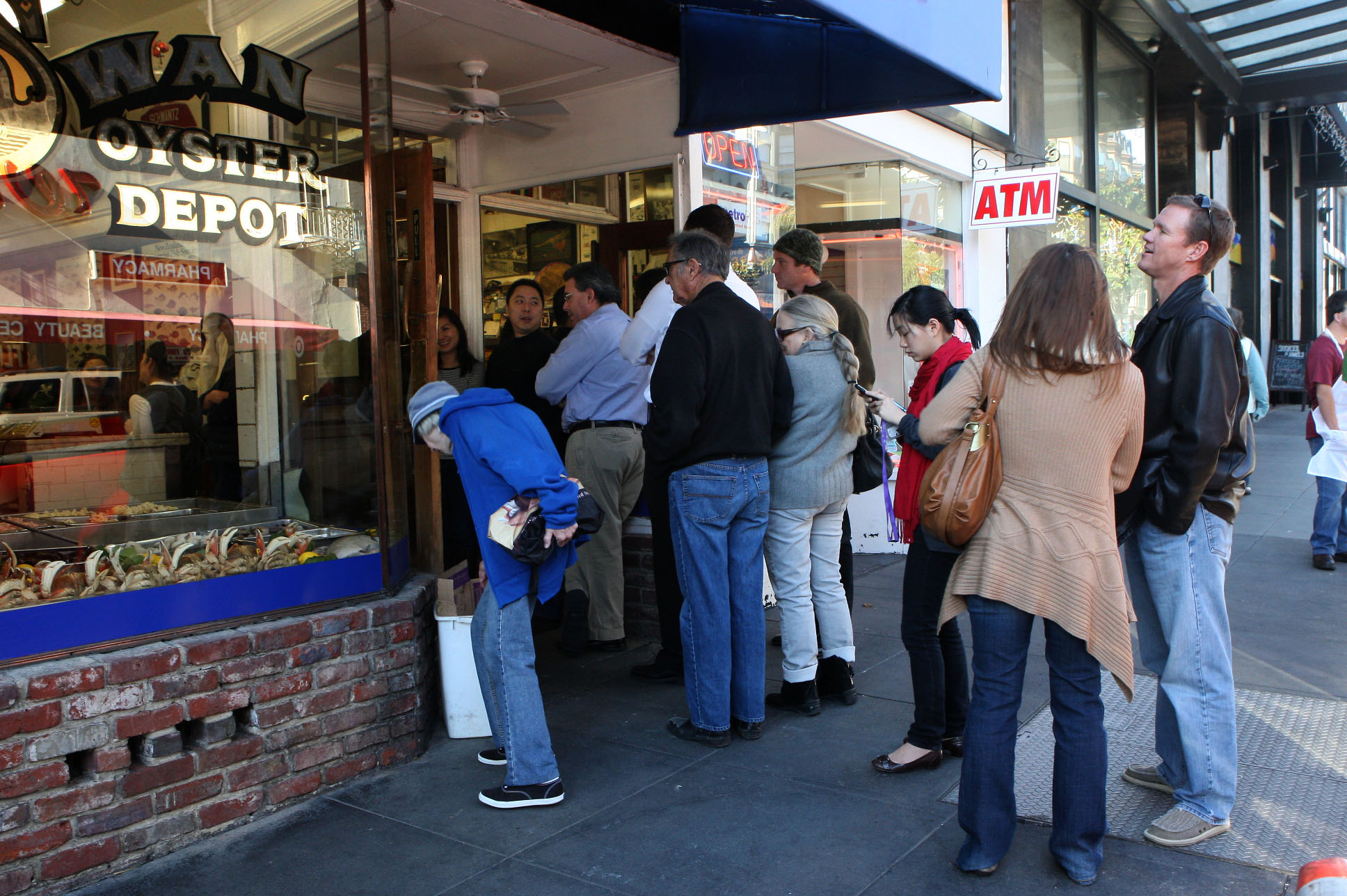 A group of people waits outside a seafood restaurant with a brick facade and a large window displaying seafood. There's an ATM sign above the entrance.