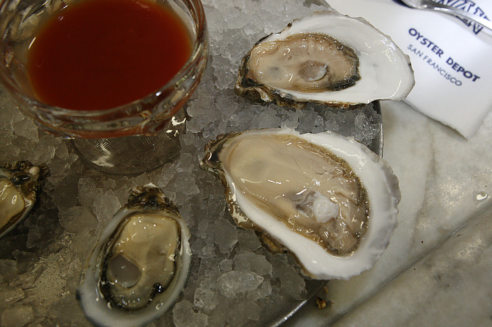 The image shows raw oysters on ice beside a glass of red sauce. A napkin labeled "Oyster Depot San Francisco" is visible in the corner.