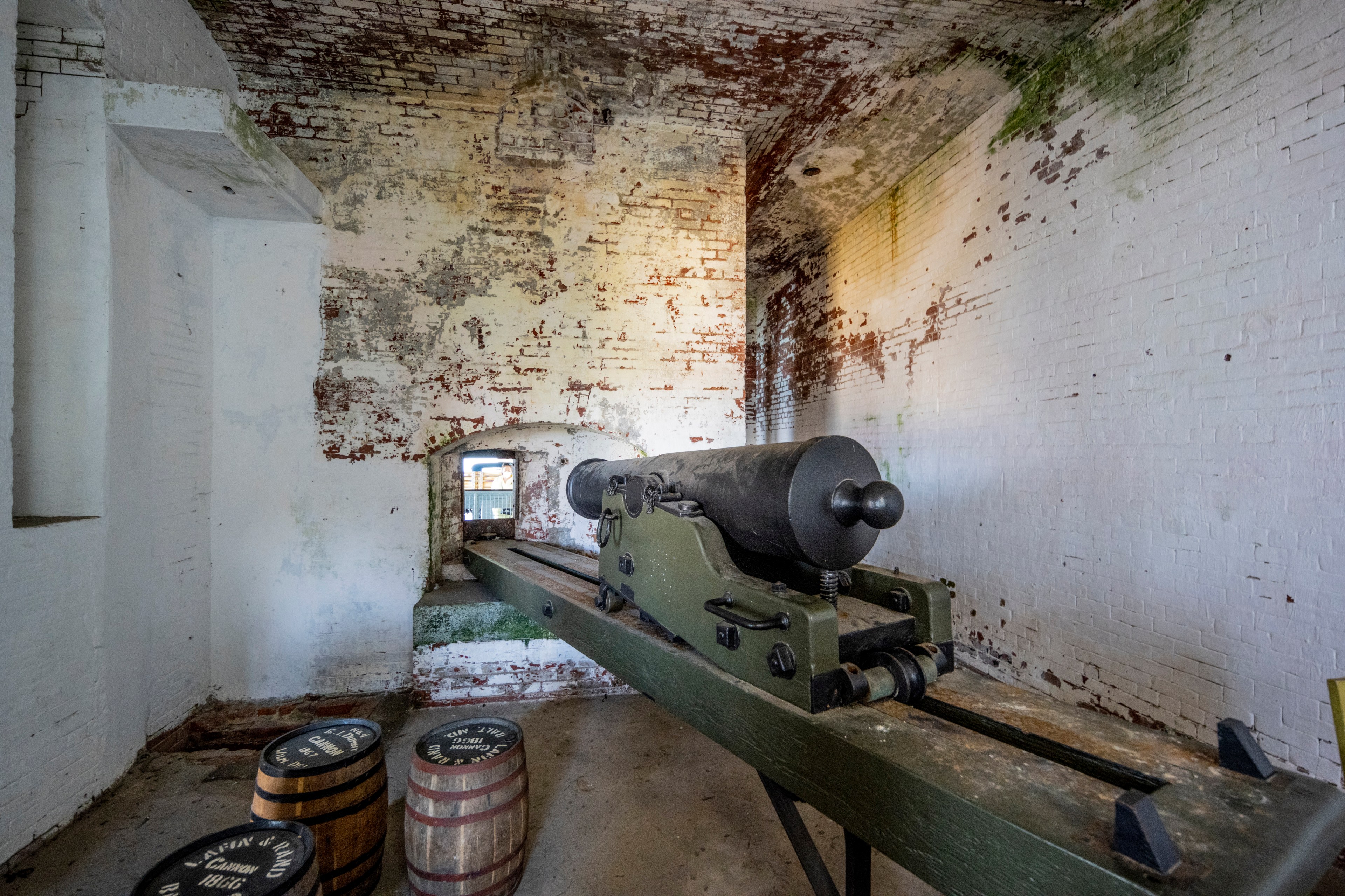The image shows an old cannon inside a rustic, brick-walled room. The walls are weathered with peeling paint. Barrels labeled “1863” are on the floor.