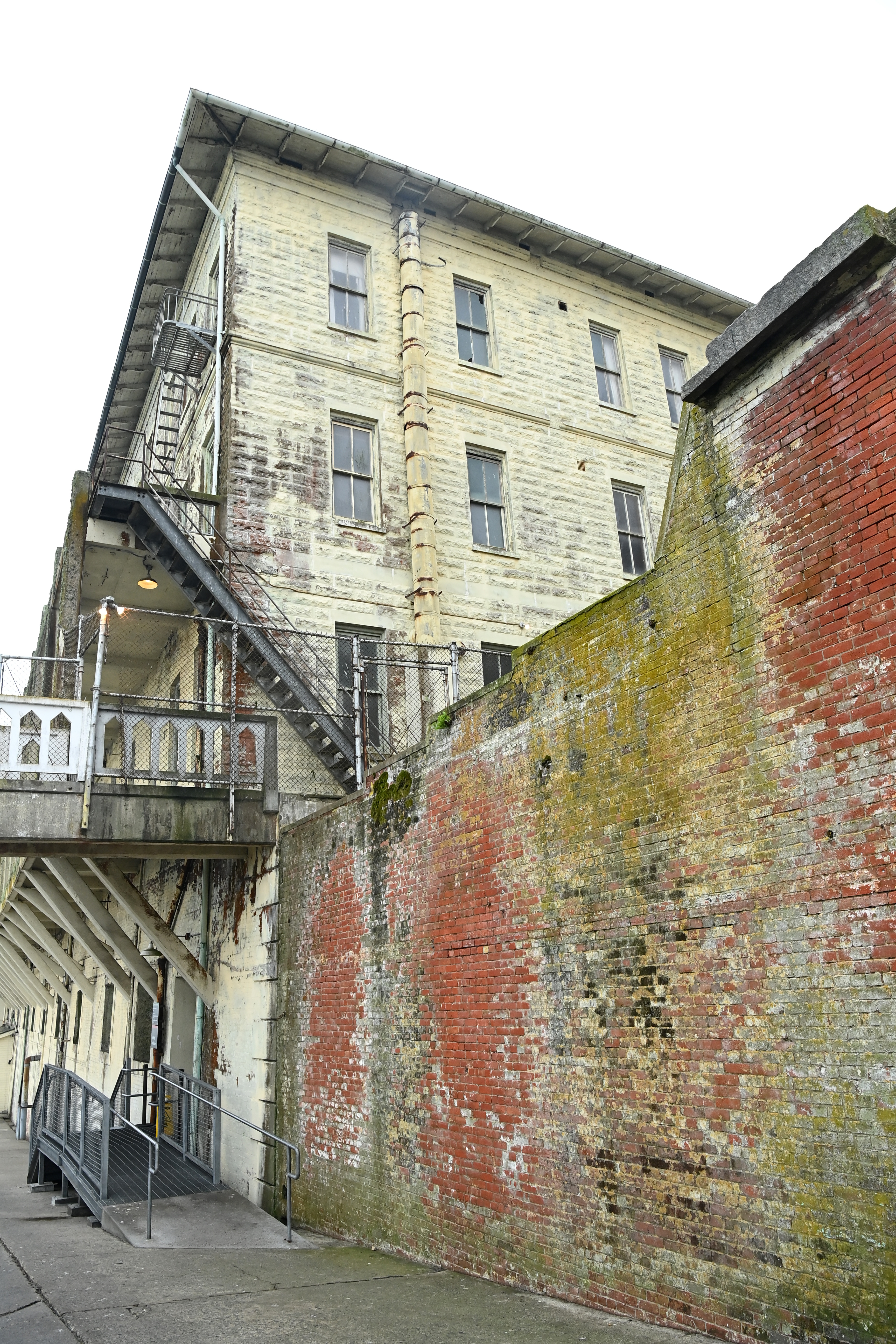 An old, weathered building with peeling paint and tall windows stands beside a moss-covered brick wall. A metal staircase and fenced walkway are visible.