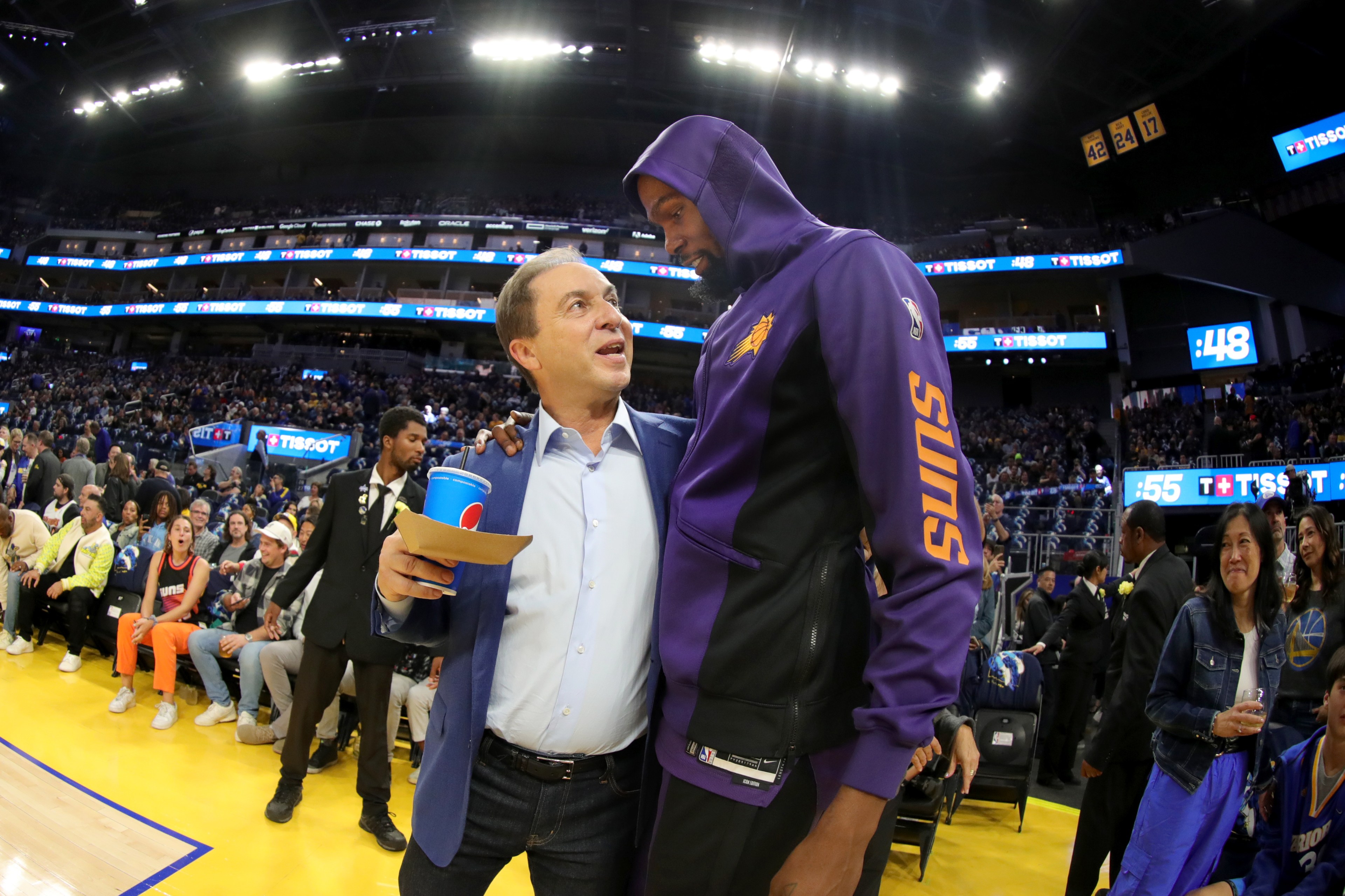 A man in a blue suit and another man in a purple "Suns" hoodie are talking and smiling on a basketball court, with a crowd and security in the background.