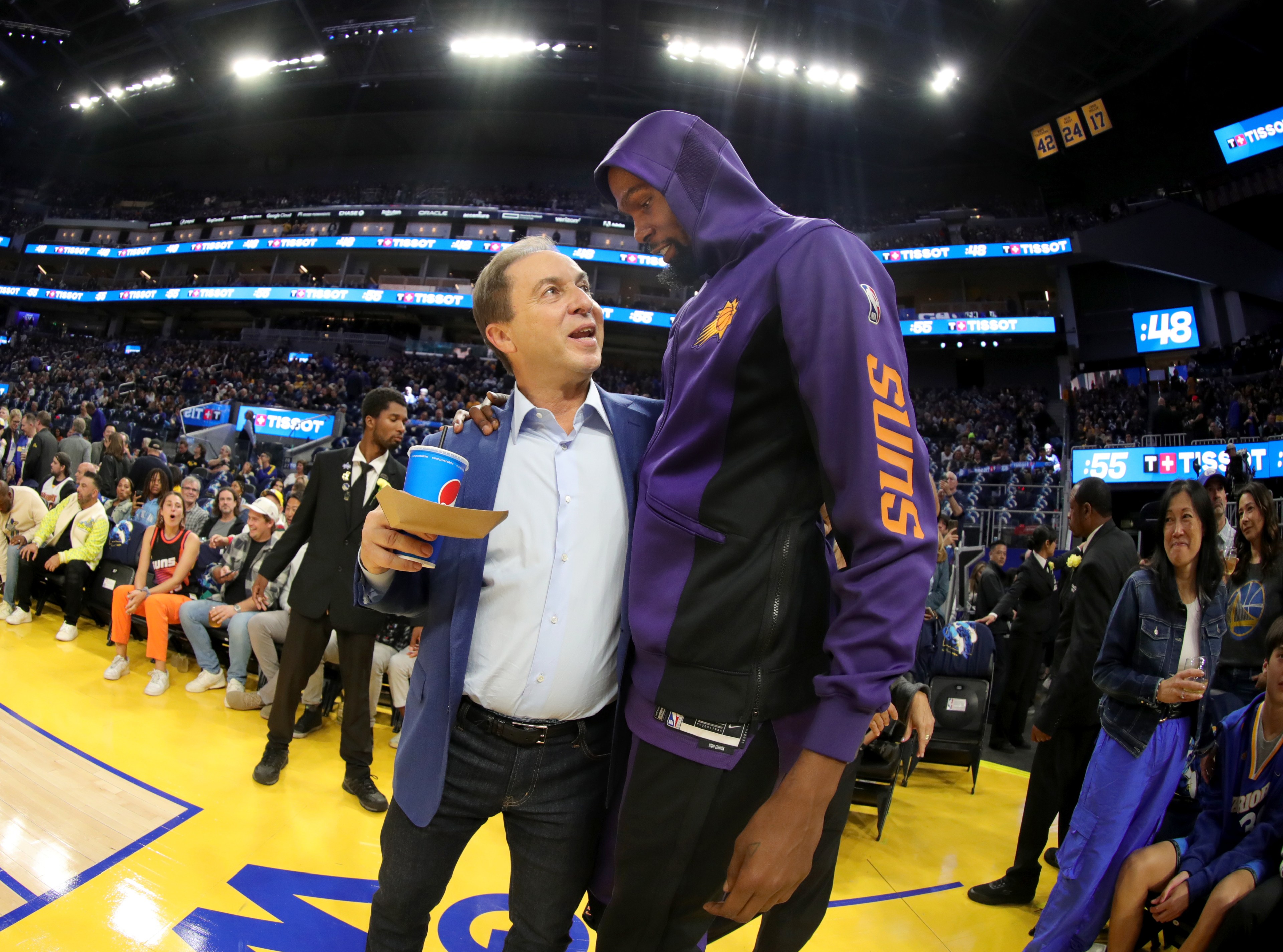 A man in a blue suit and another man in a purple "Suns" hoodie are talking and smiling on a basketball court, with a crowd and security in the background.