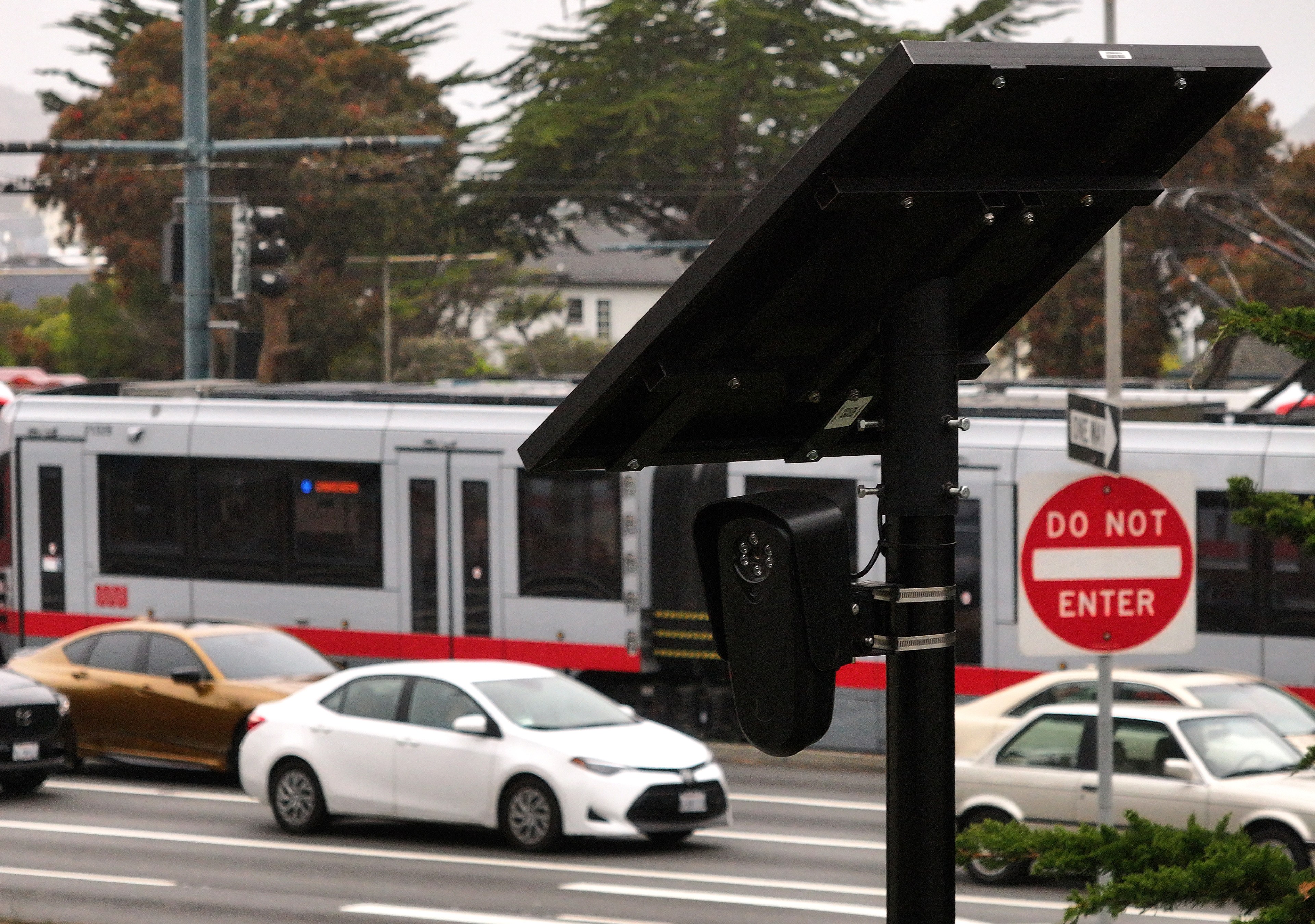 The image shows a street scene with a "Do Not Enter" sign, several cars, and a moving tram. A pole with a solar panel and security camera is prominent.