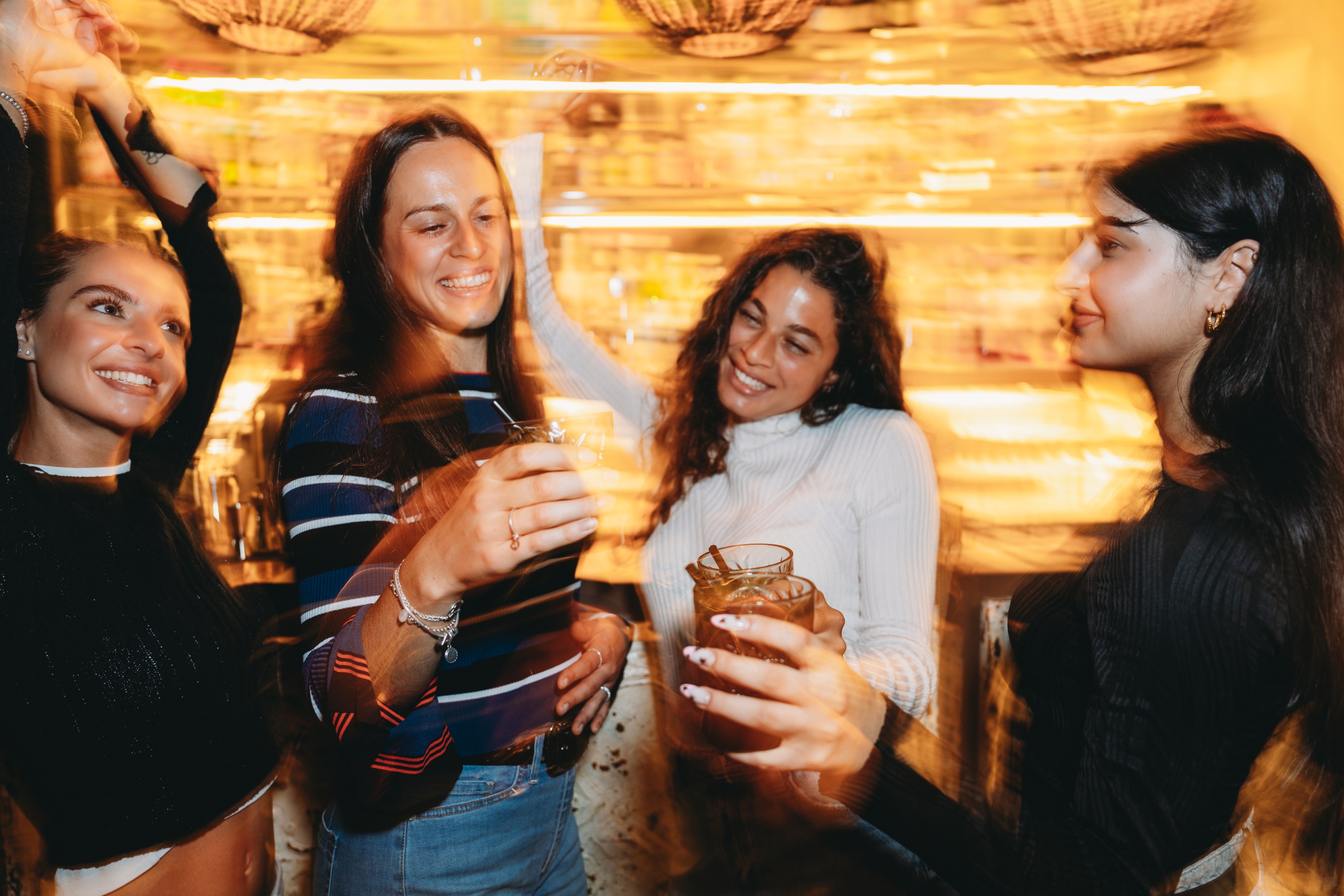 Four women are joyfully toasting with drinks in their hands at a lively bar, surrounded by warm, blurred lights, creating a festive atmosphere.