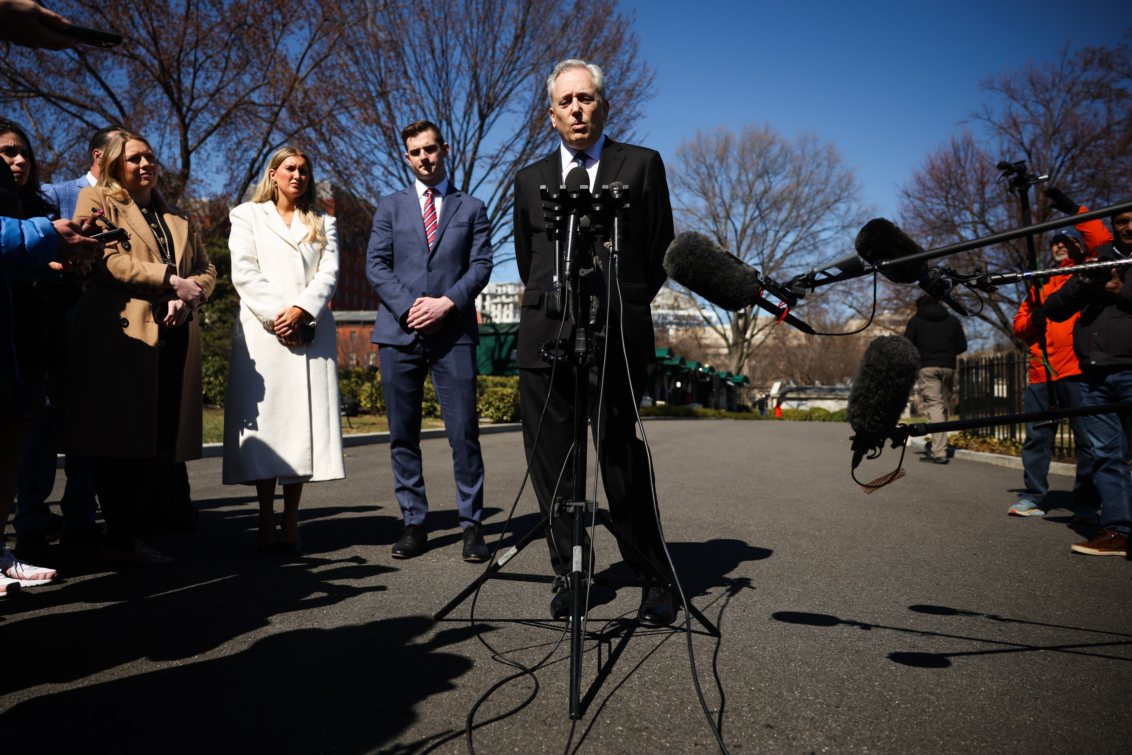 A person stands in front of multiple microphones, speaking outdoors with a clear sky. Several individuals in formal attire listen, surrounded by trees.