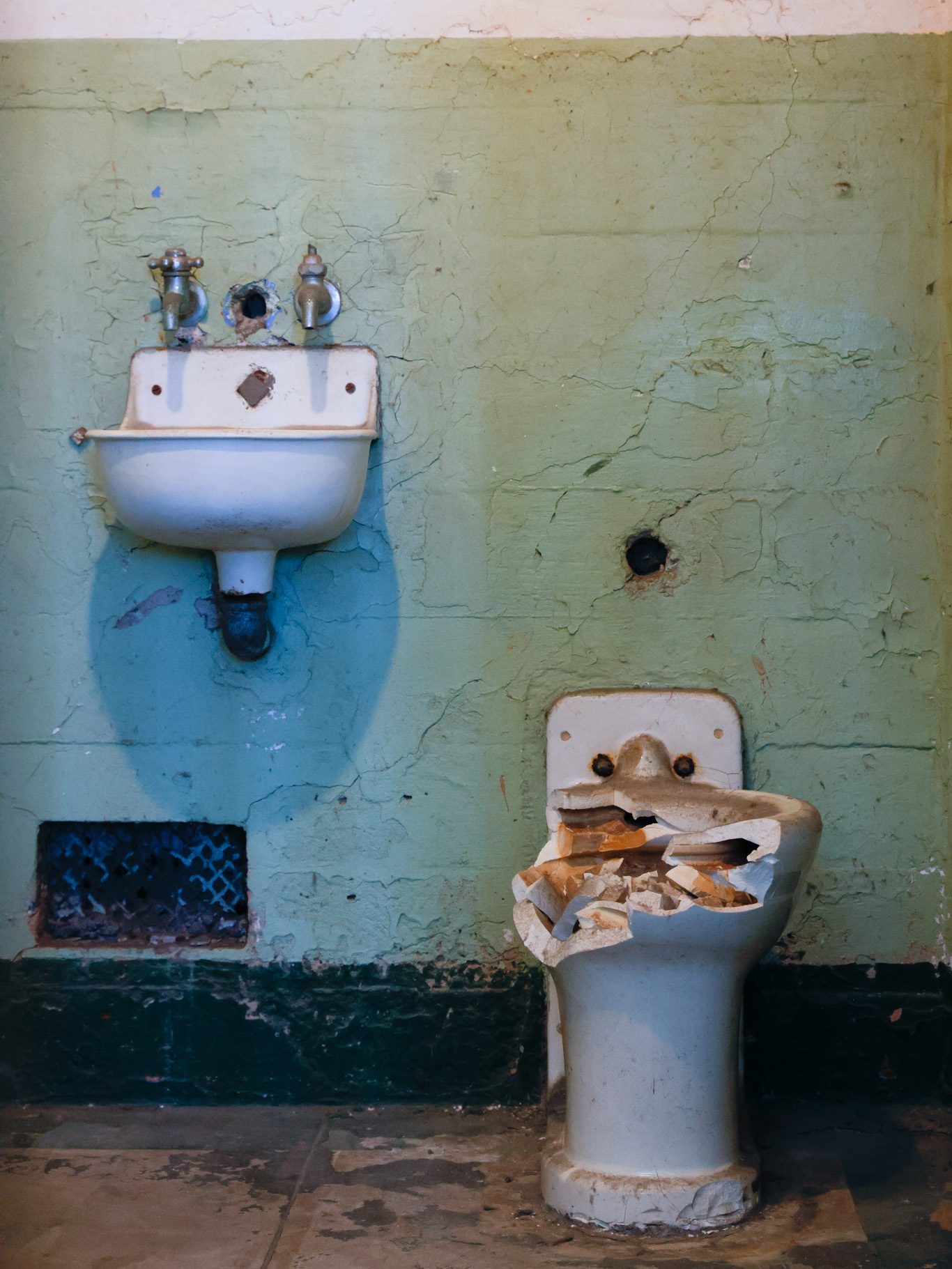 The image shows an old, discolored wall with a rusty sink and a broken toilet fixture beneath it. The wall and floor have visible wear and cracks.