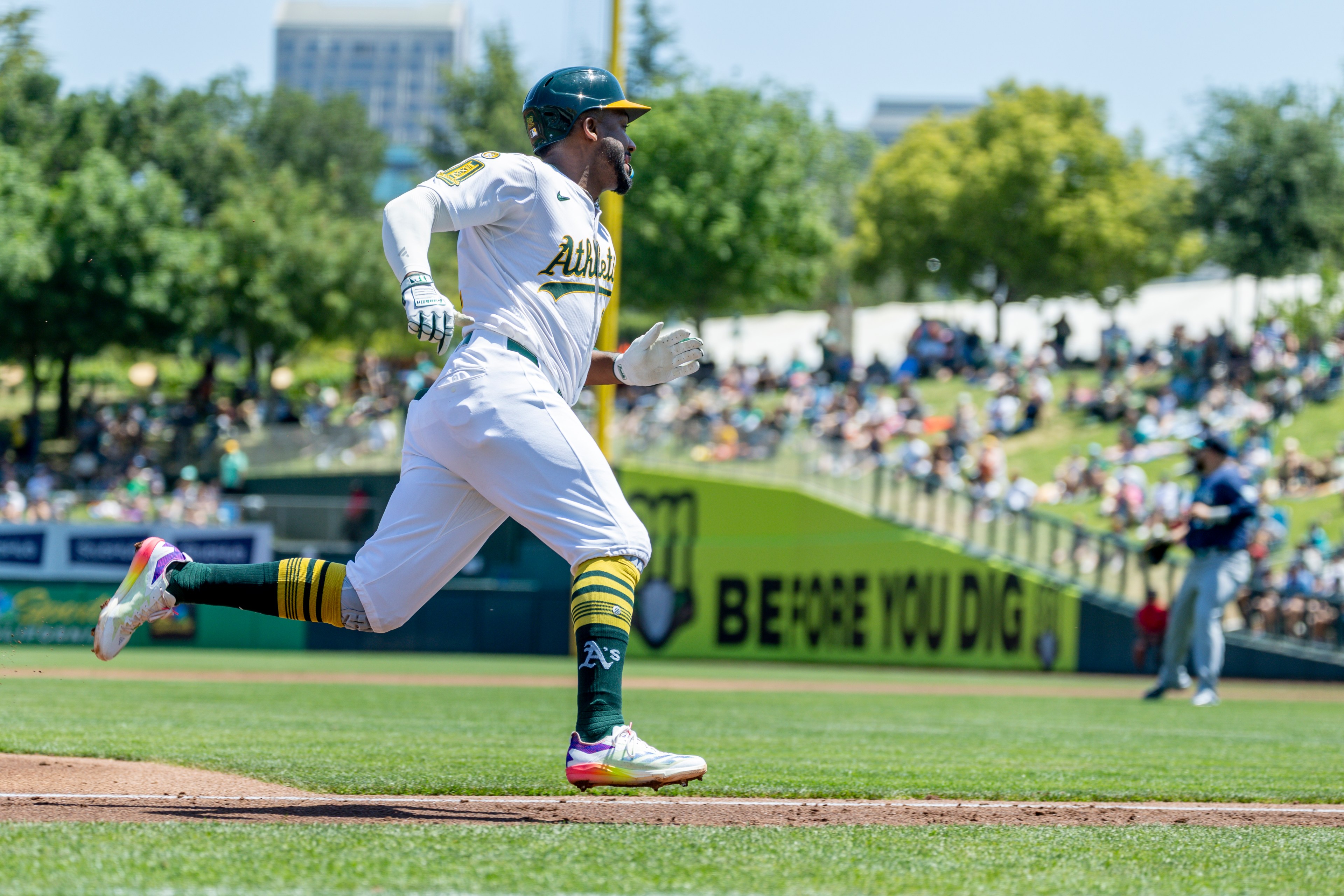 A baseball player in a white uniform and helmet is running on the field. The background shows an audience sitting on grassy slopes under trees.