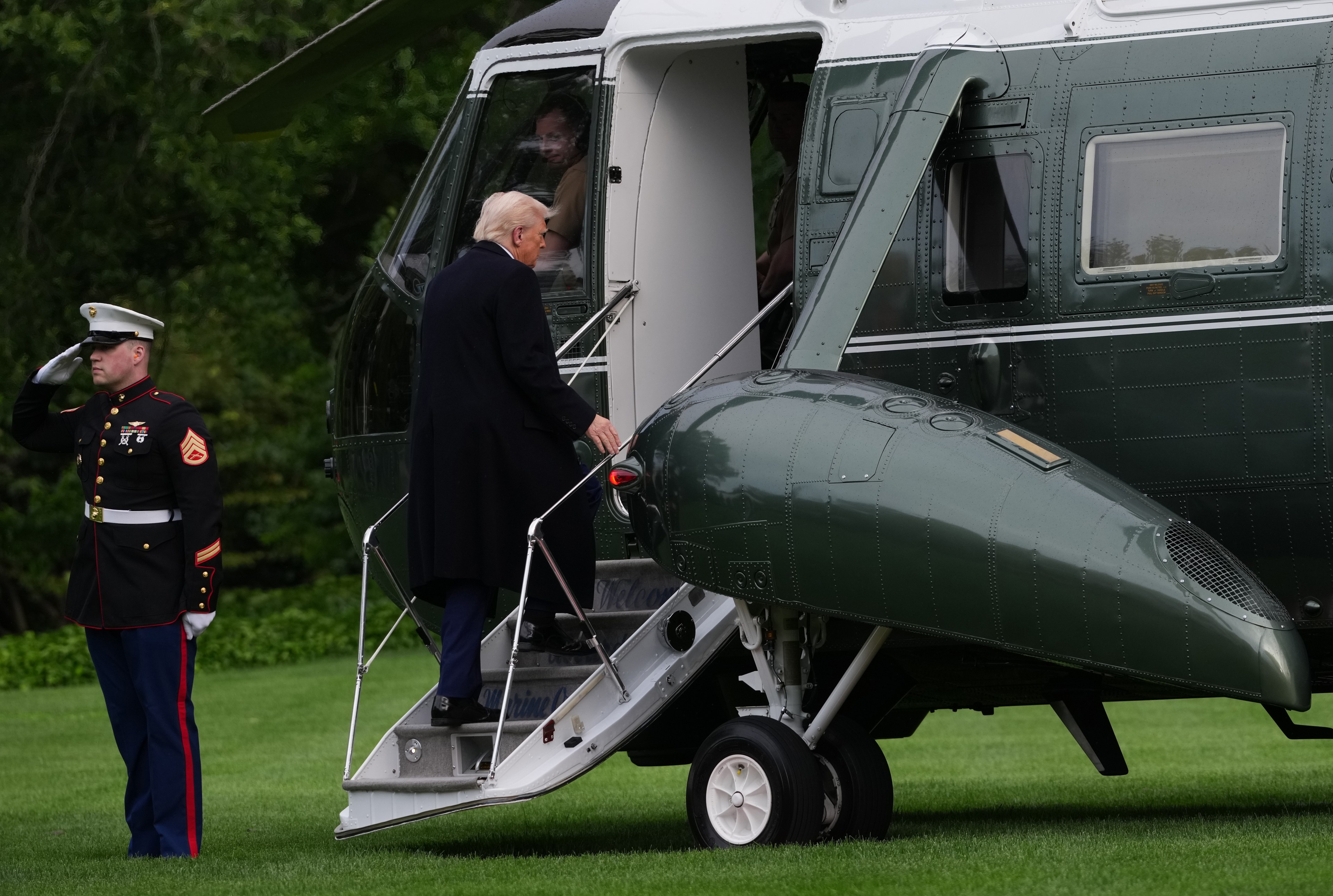 A man in a dark coat is boarding a green and white helicopter. A uniformed Marine stands saluting nearby. The scene is set on a grassy area.