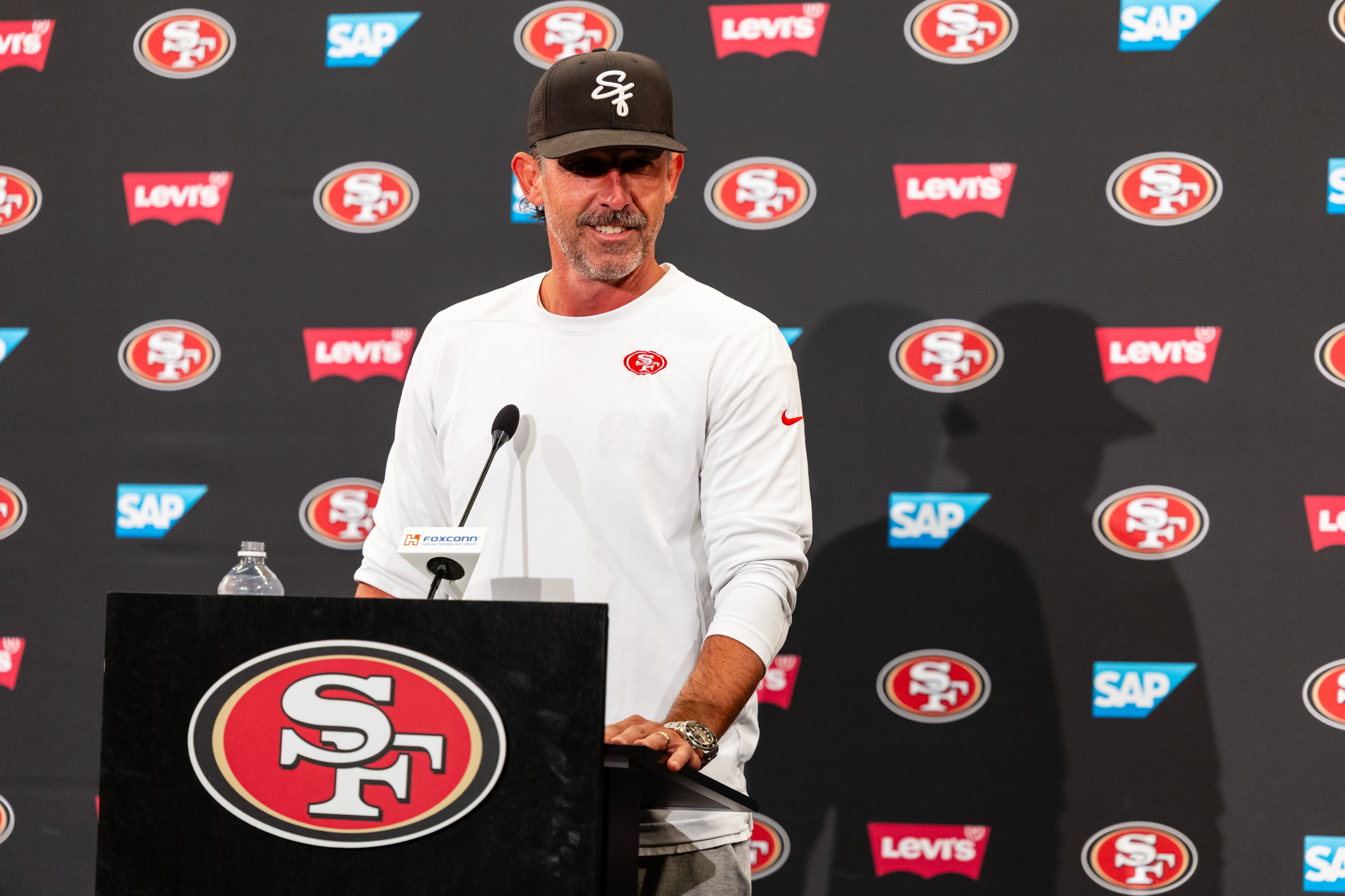 A man in a white long-sleeve shirt and black cap stands at a podium with an "SF" logo, against a backdrop featuring the same logo and sponsor names.