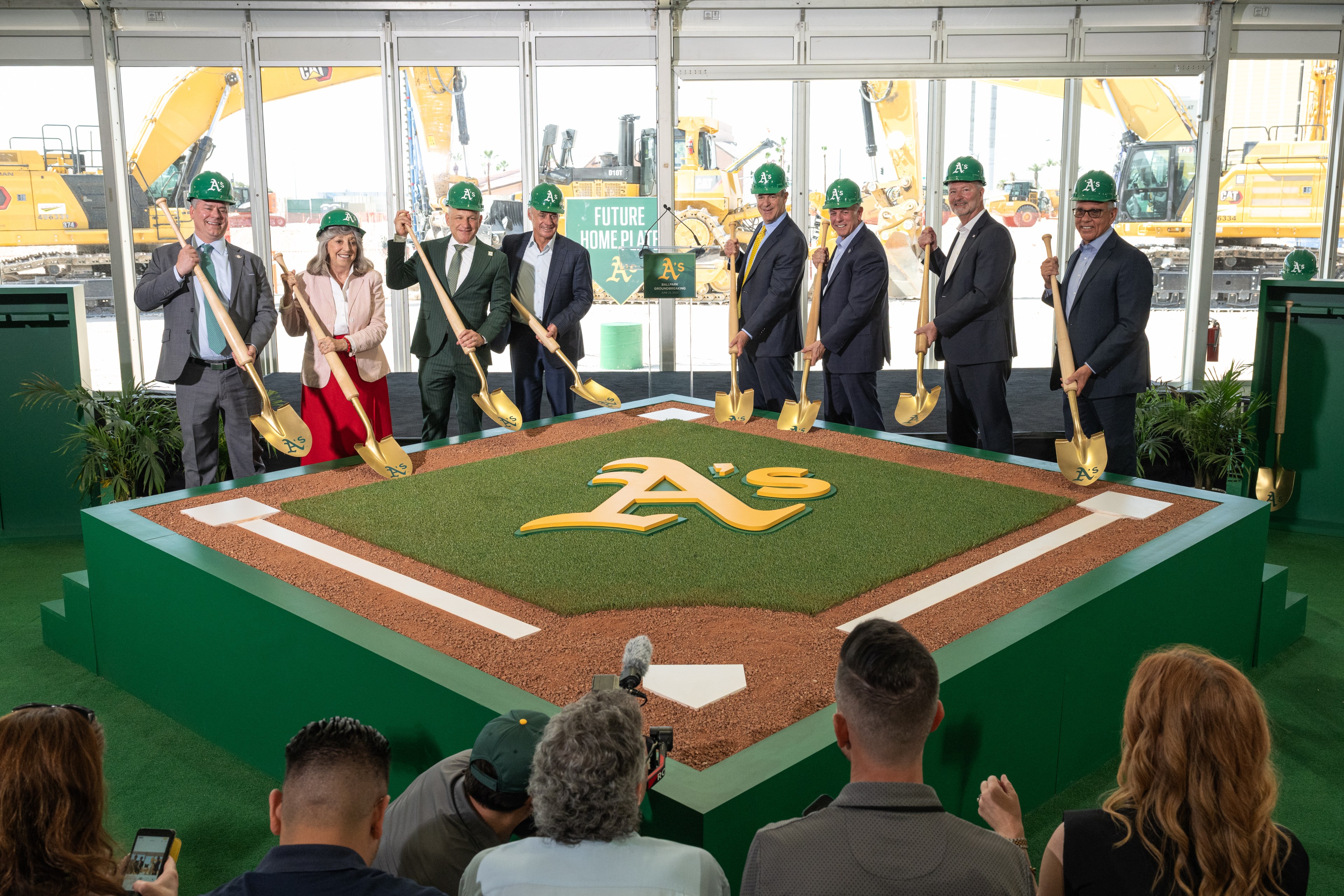 Nine people wearing green hard hats stand smiling, holding golden shovels over a mock baseball home plate area with an "A's" logo, during a groundbreaking event.