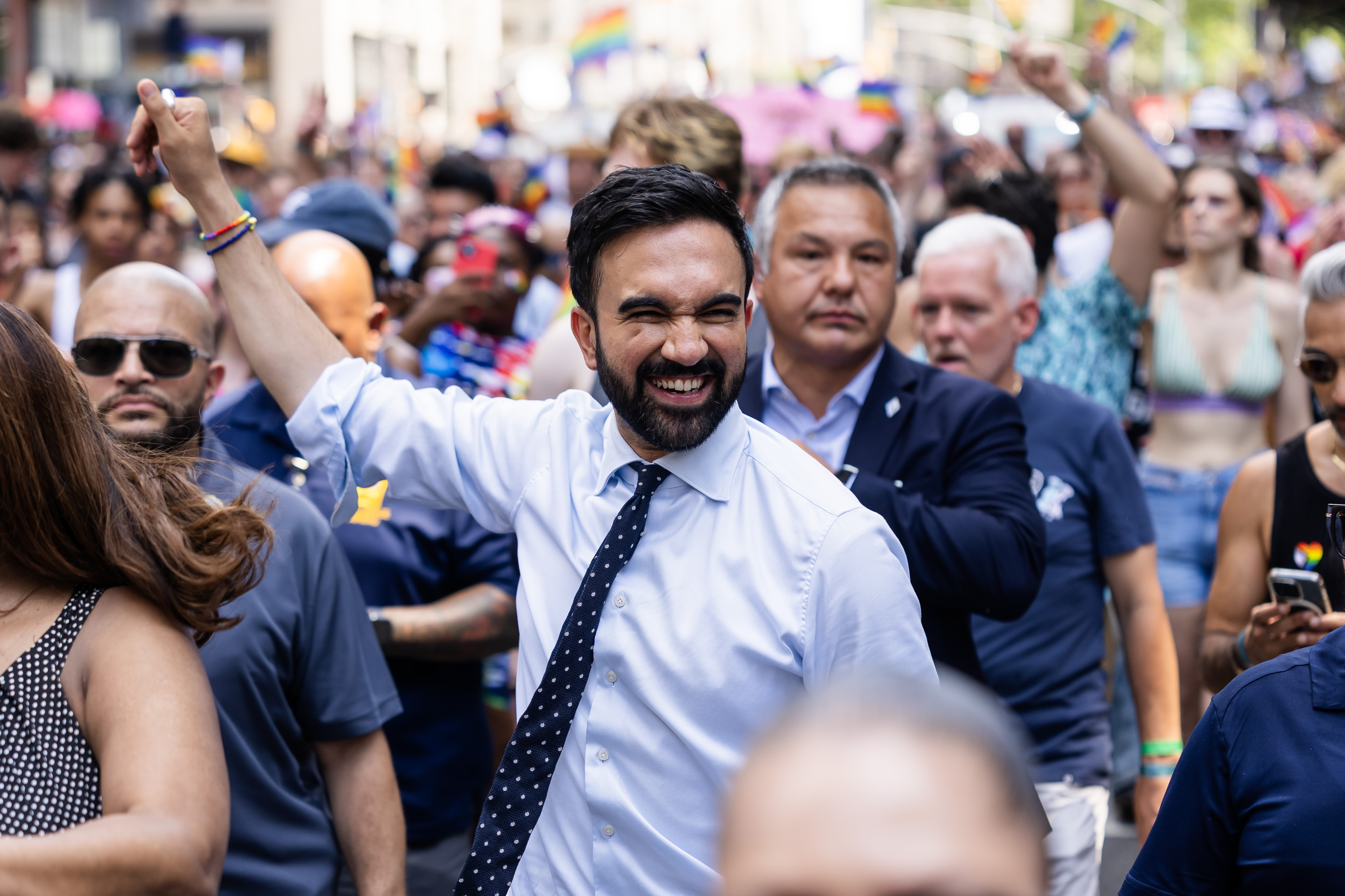 A group of people are joyfully marching in a parade. The central figure is smiling, wearing a white shirt and tie, with raised fists in a crowded street.