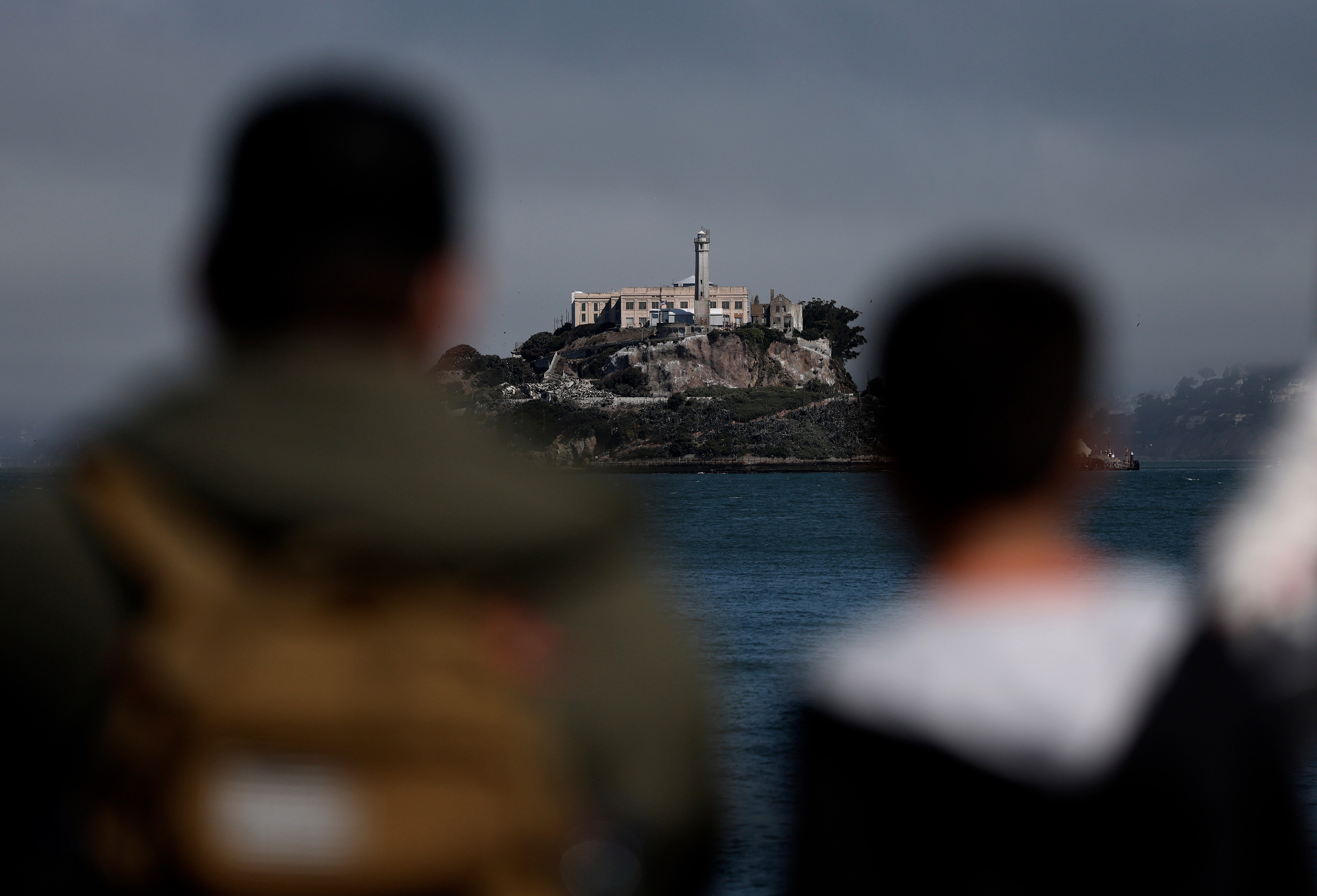 Two blurred figures gaze towards a distant island with a prominent building and lighthouse. The scene is set against a cloudy sky and calm water.