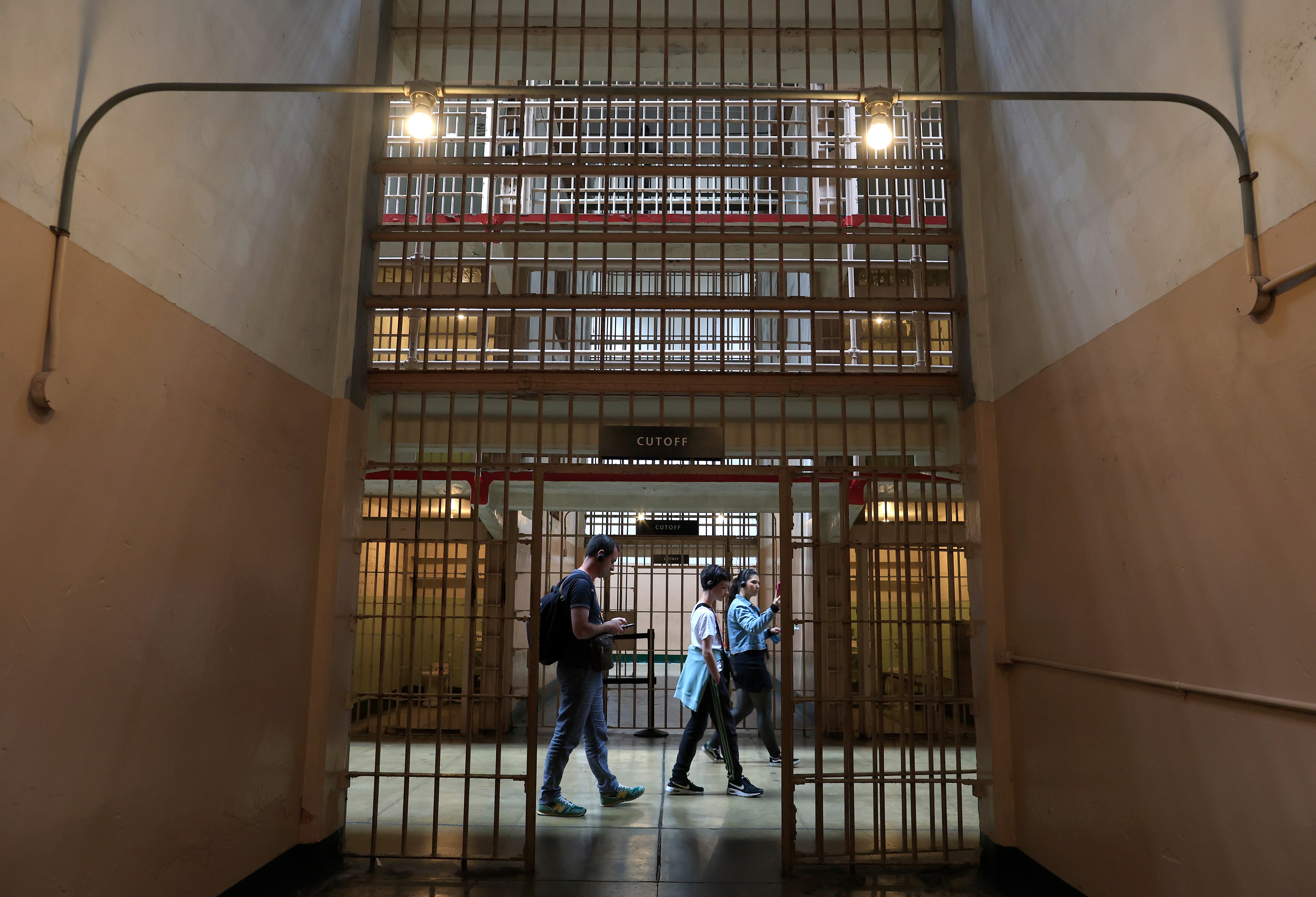 Three people walk through a dimly lit corridor with metal bars surrounding them, resembling a prison interior marked &quot;CUTOFF.&quot; Two overhead lights illuminate the space.
