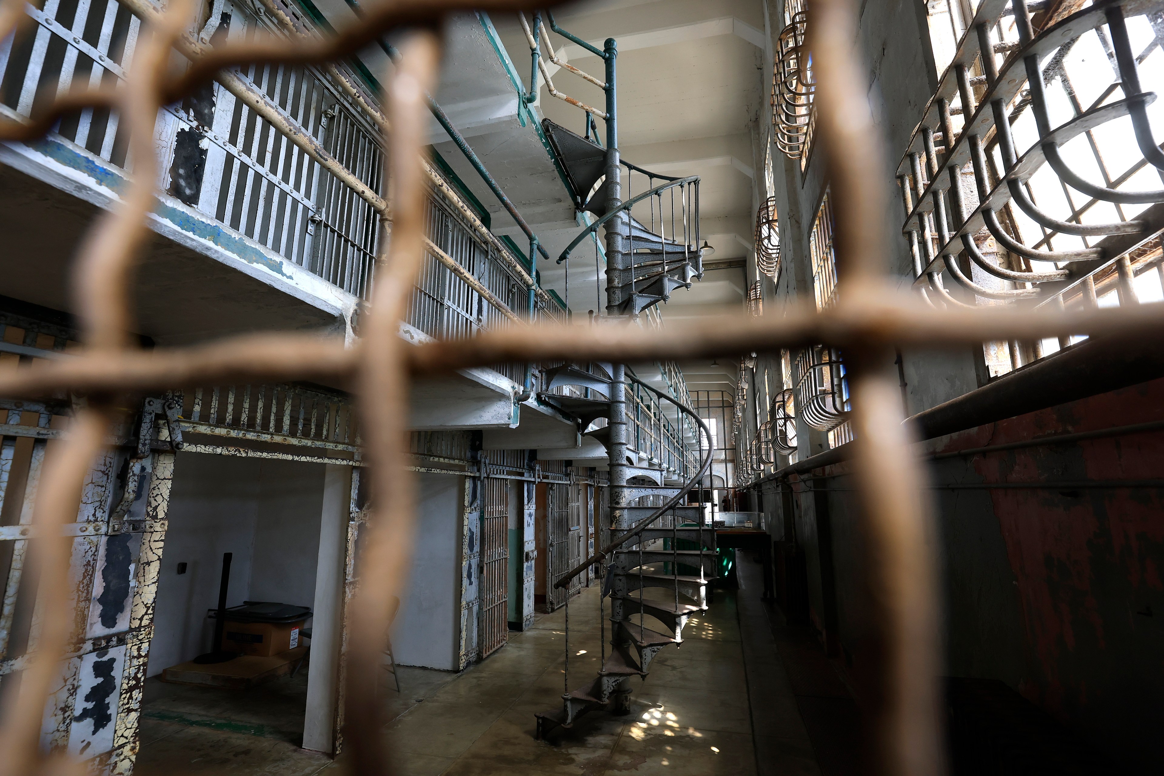 The image shows the interior of an old prison with a spiral staircase, barred windows, and two levels of cells. The scene is viewed through a rusted wire fence.