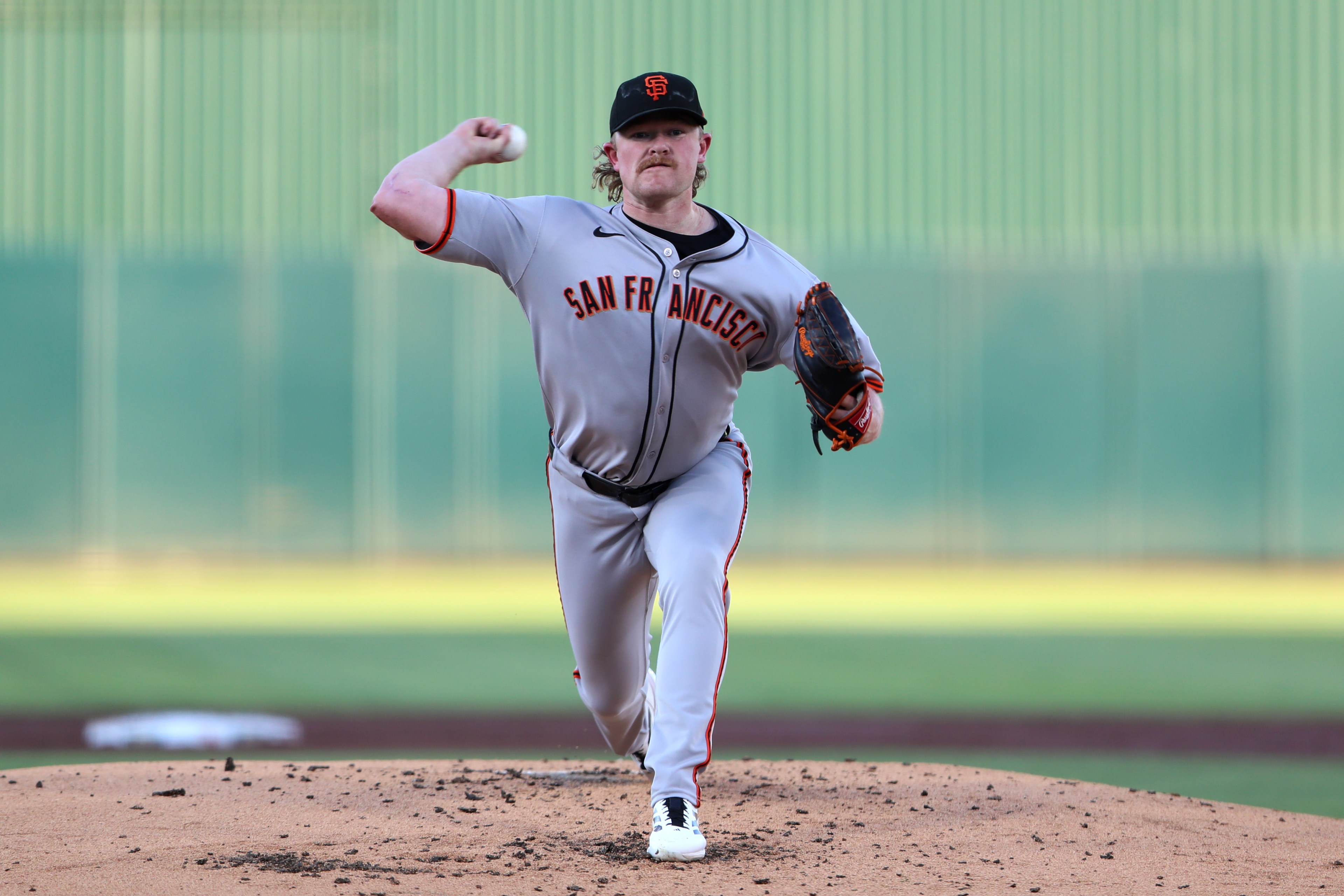 A baseball pitcher in a gray "San Francisco" uniform is mid-throw on a mound, wearing a black cap. The outfield wall is blurred in the background.