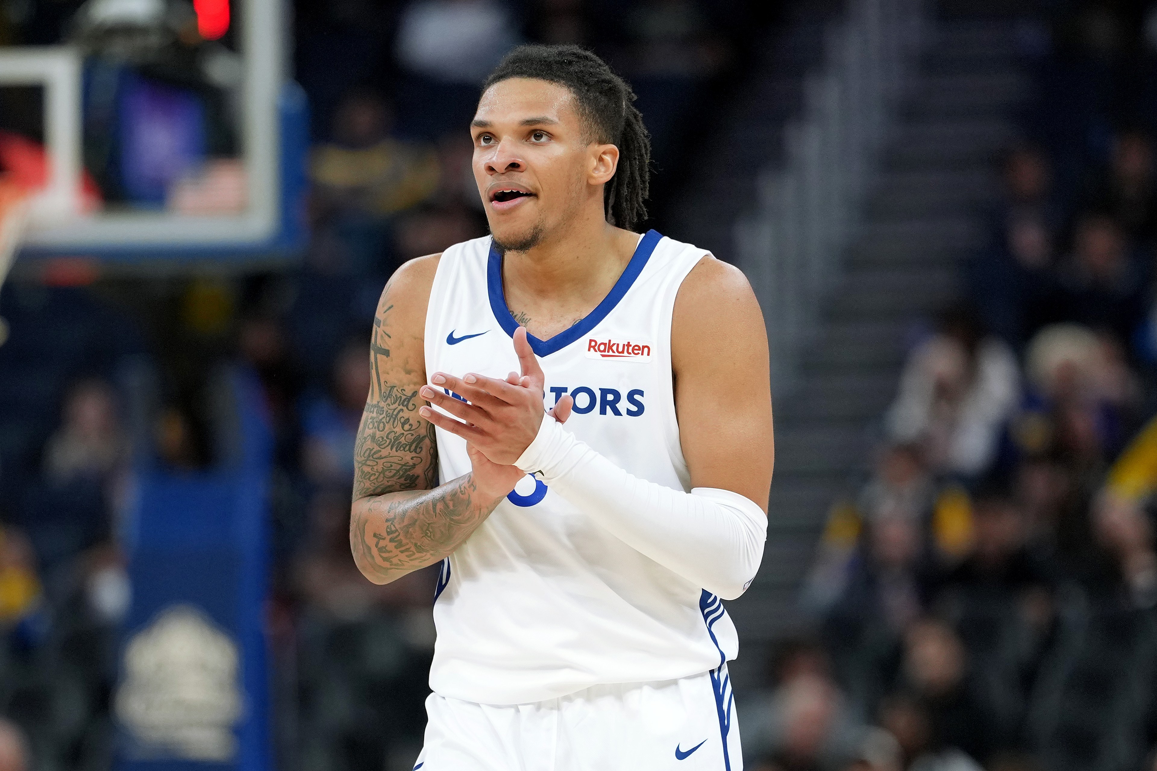 A basketball player in a white jersey with blue trim is clapping on the court. The blurred audience is in the background.