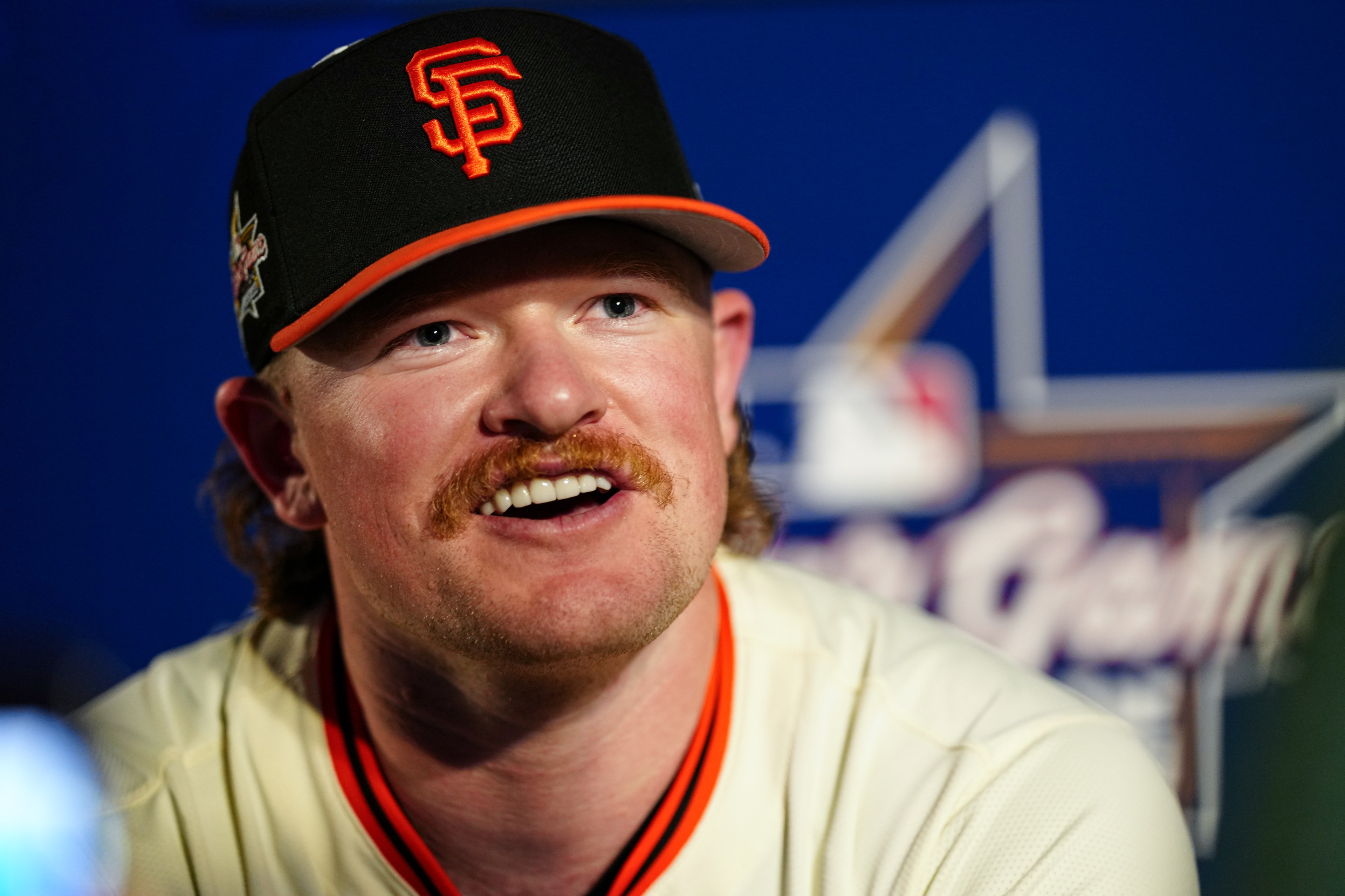 A man with a mustache wears a black SF cap and a baseball jersey. He is smiling and looking slightly upwards, with a blurred sporting event logo behind him.