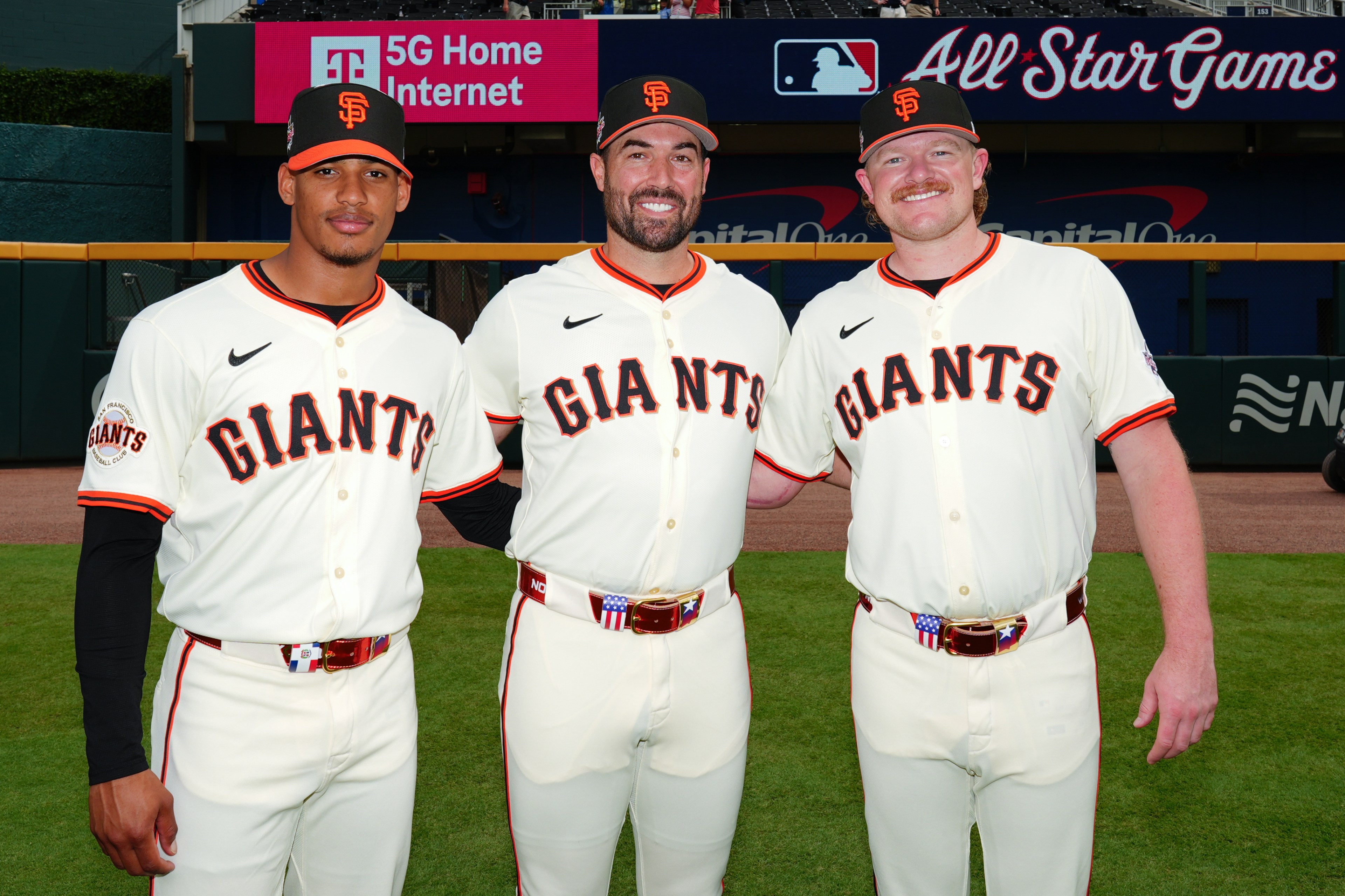 Three baseball players in Giants uniforms stand on a field, smiling with arms around each other. They wear hats and have a backdrop of All-Star Game signage.