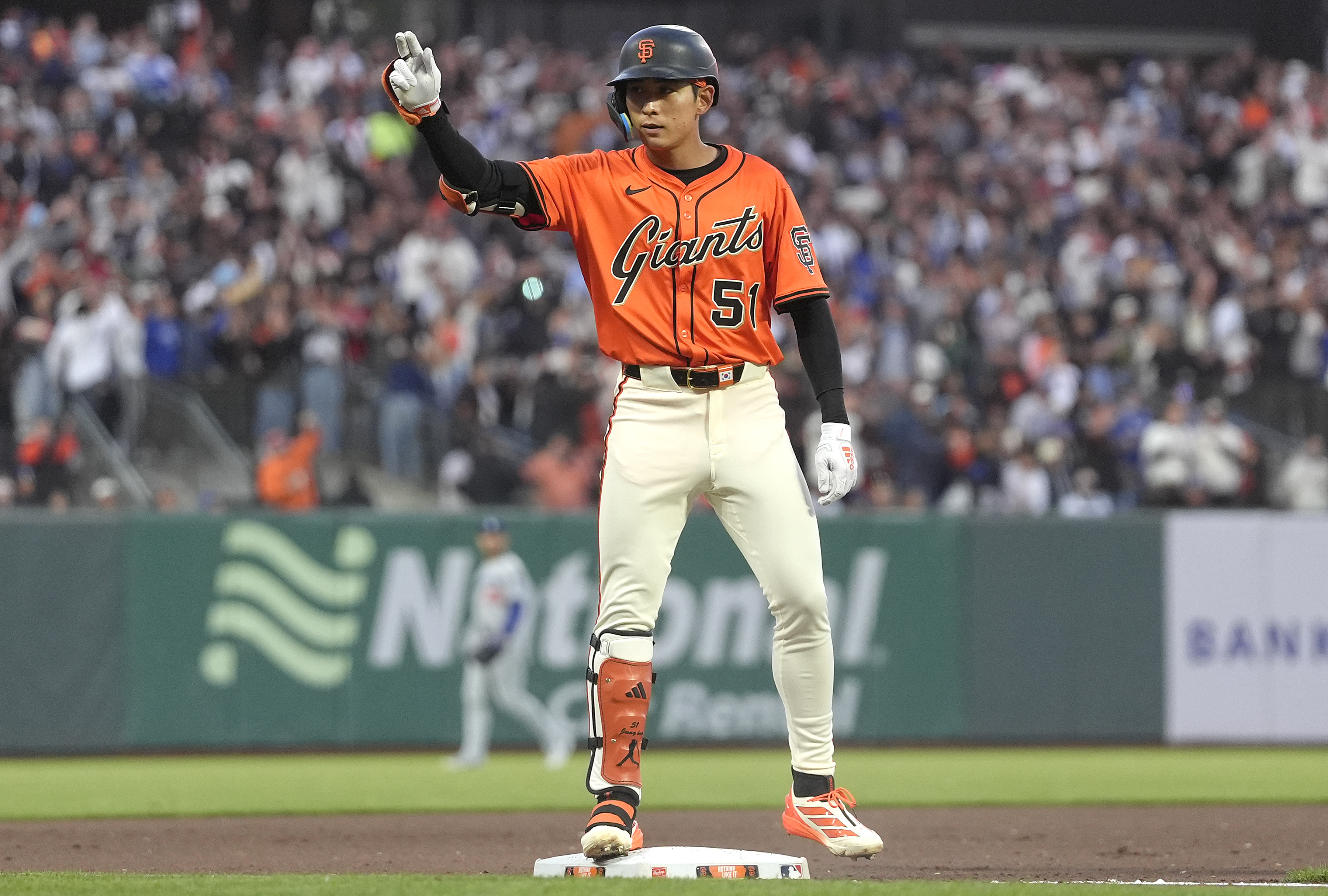 A baseball player wearing an orange Giants jersey stands on a base, gesturing with one hand up. The background shows a crowd and field signage.