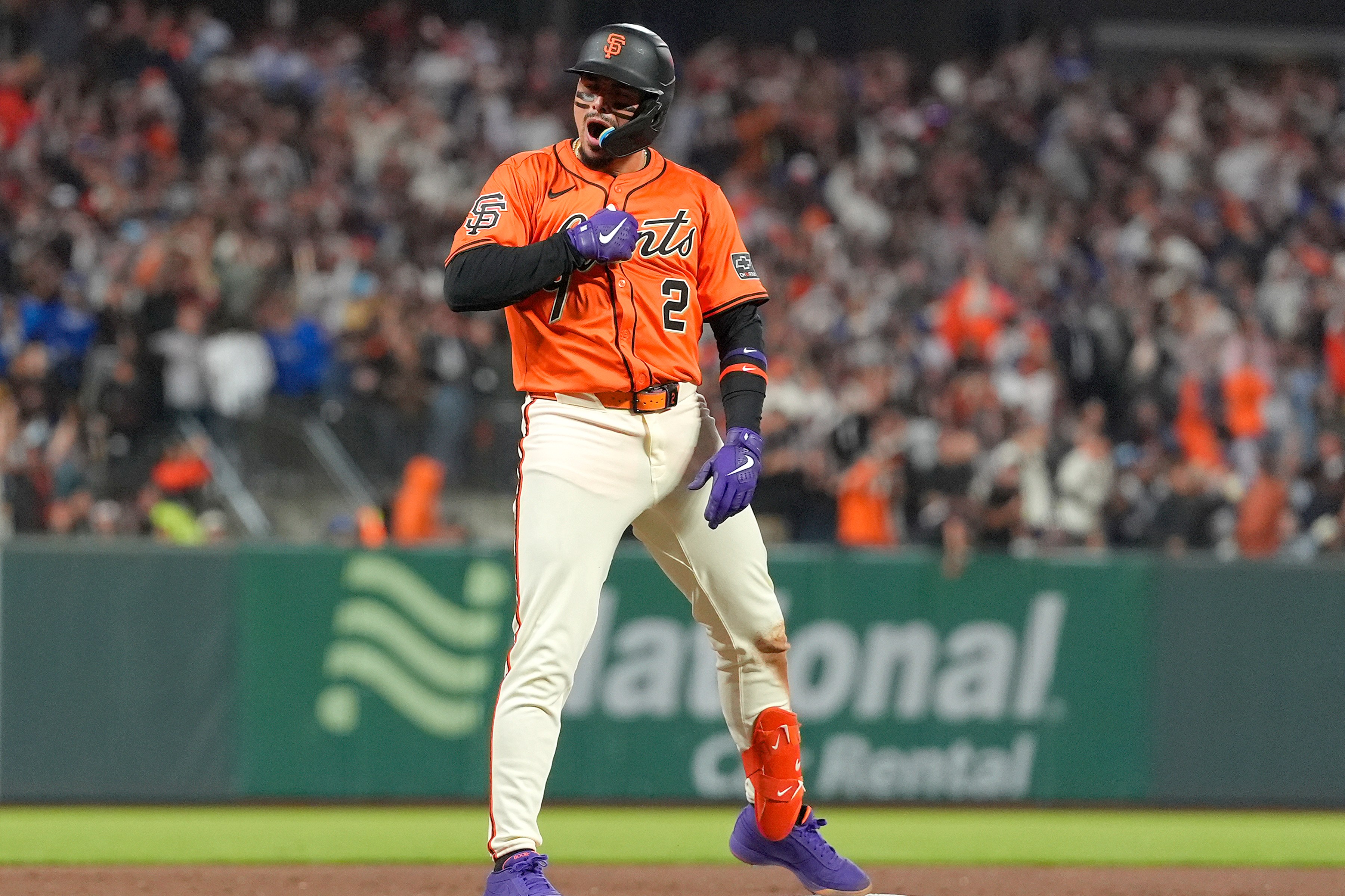 A baseball player in an orange jersey and white pants celebrates on a base. He wears a helmet, purple gloves, and bright orange protective gear amid a cheering crowd.