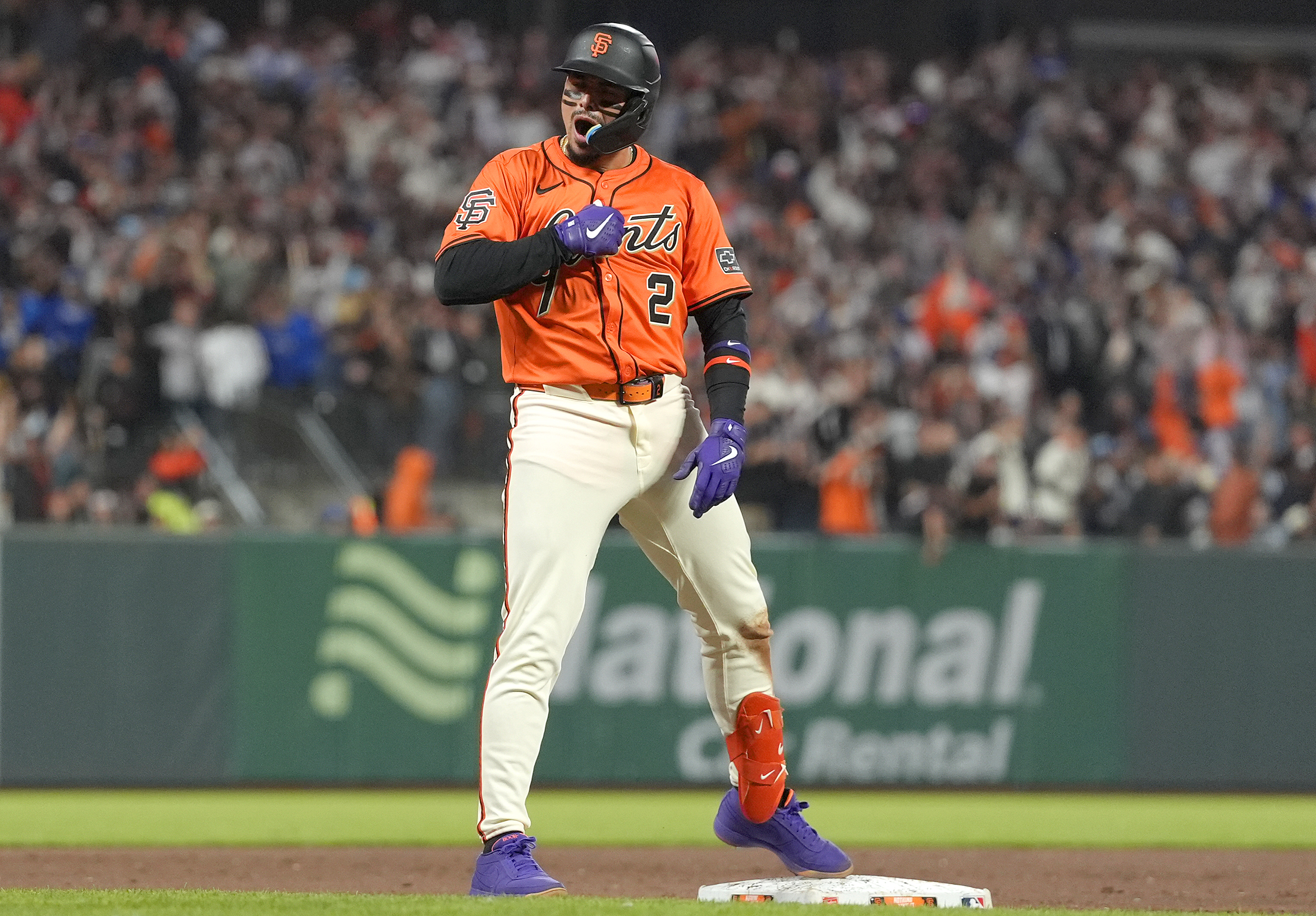 A baseball player in an orange jersey and white pants celebrates on a base. He wears a helmet, purple gloves, and bright orange protective gear amid a cheering crowd.