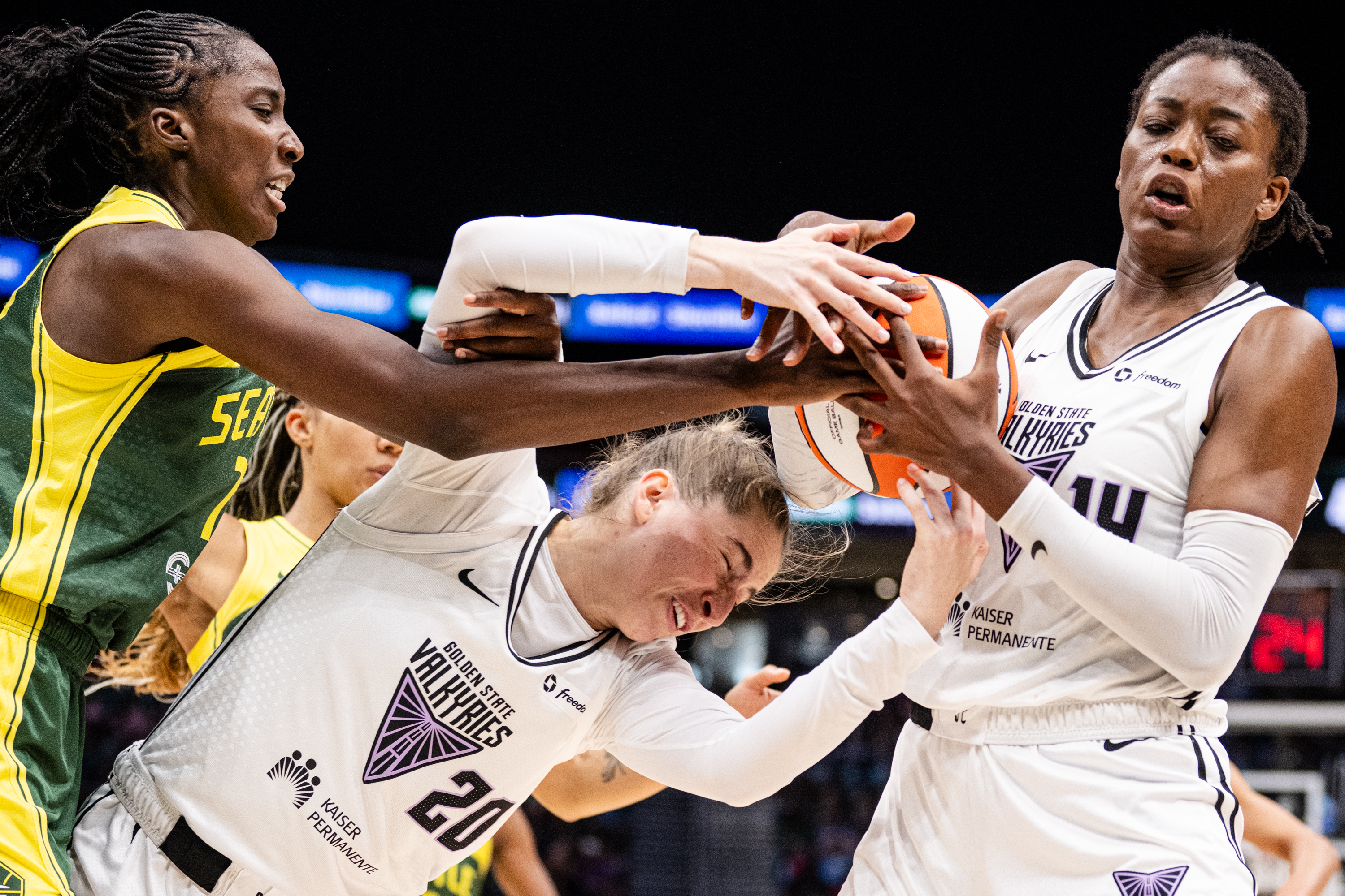 Three basketball players are vying intensely for control of the ball. Two players in white jerseys and one in a green jersey are in a tense struggle.