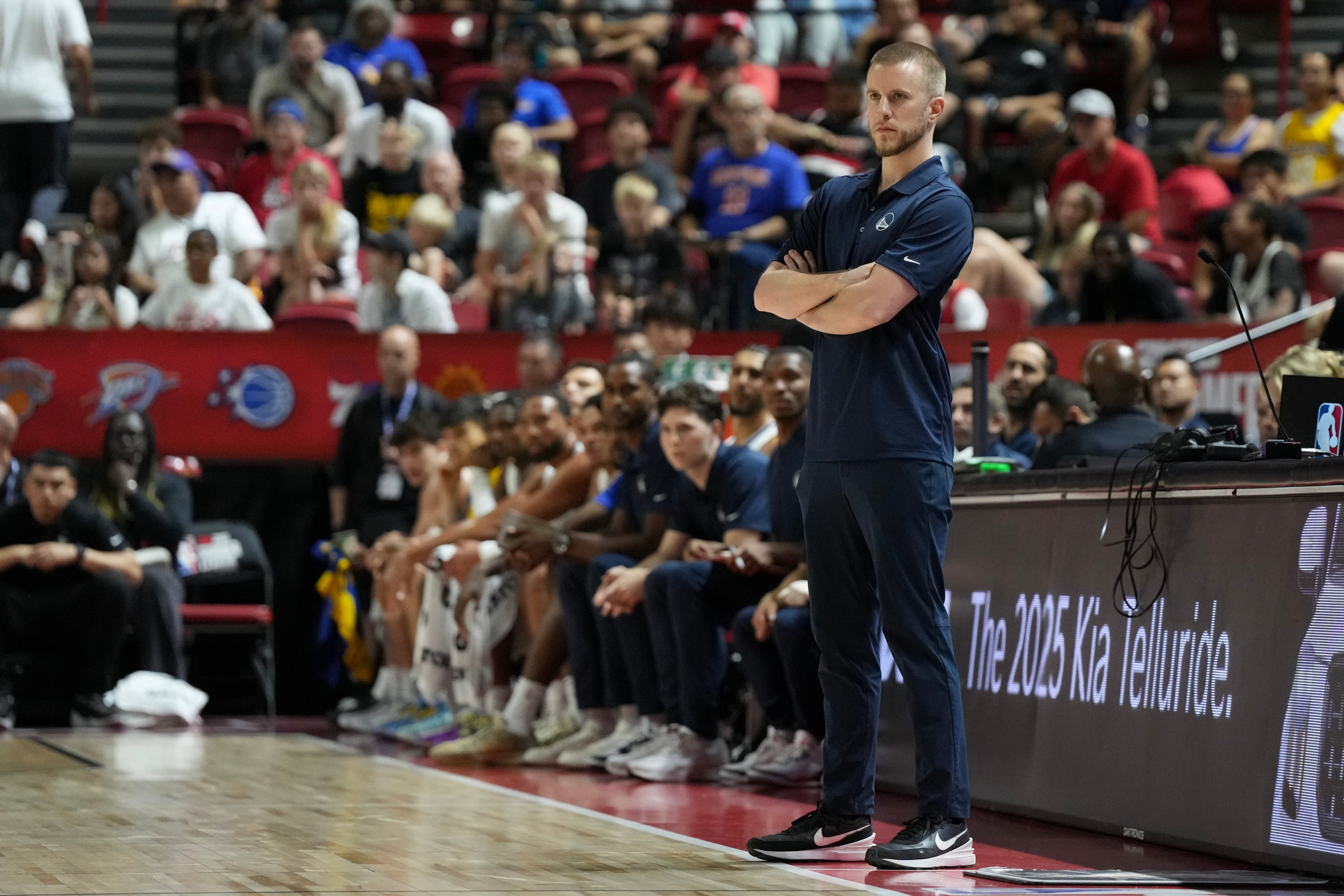 A coach stands with arms crossed on a basketball court's sideline, while players sit on the bench and an audience watches intently.
