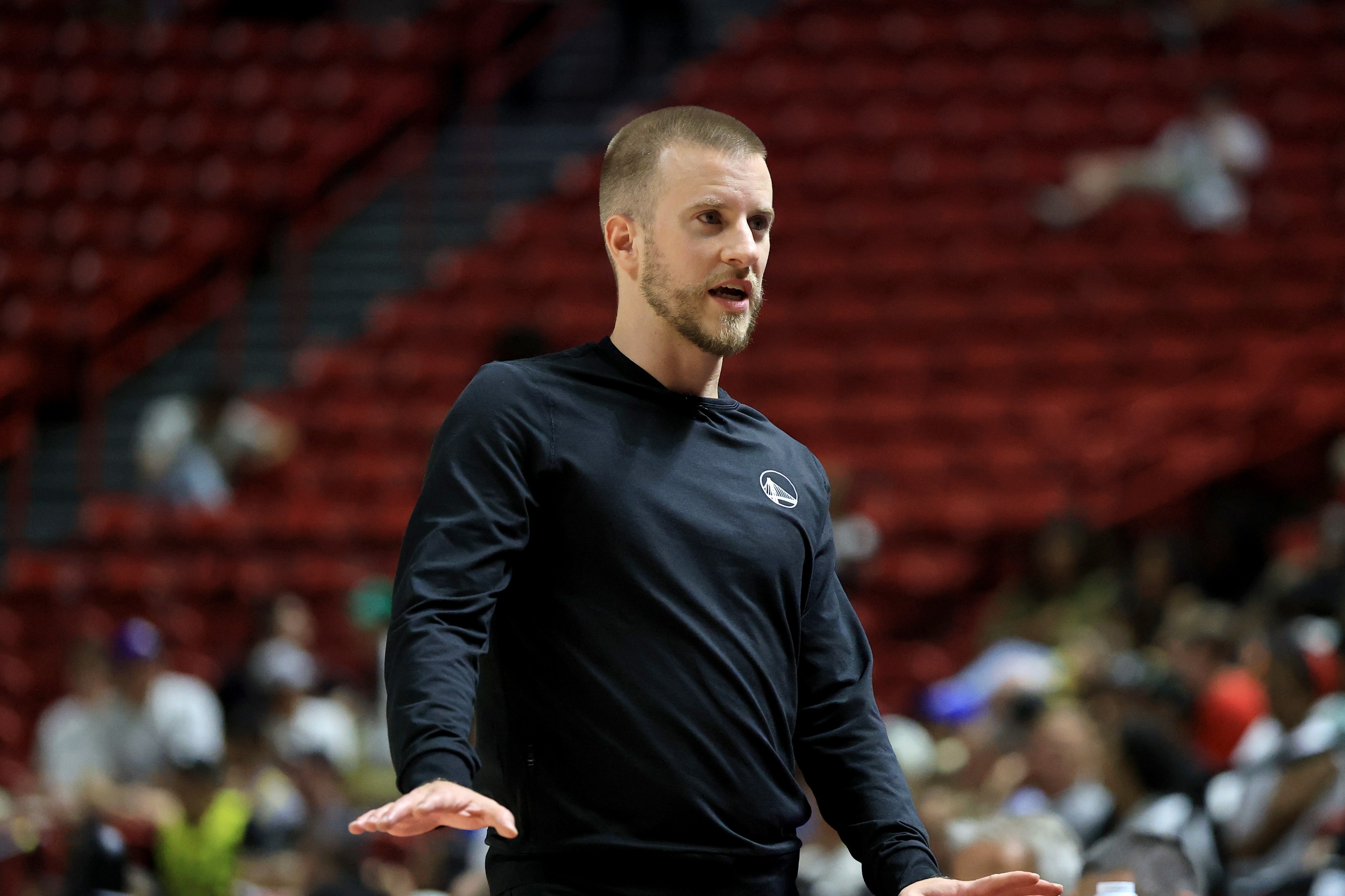 A man in a black long-sleeve shirt is speaking passionately in an indoor setting with blurred spectators in the background.