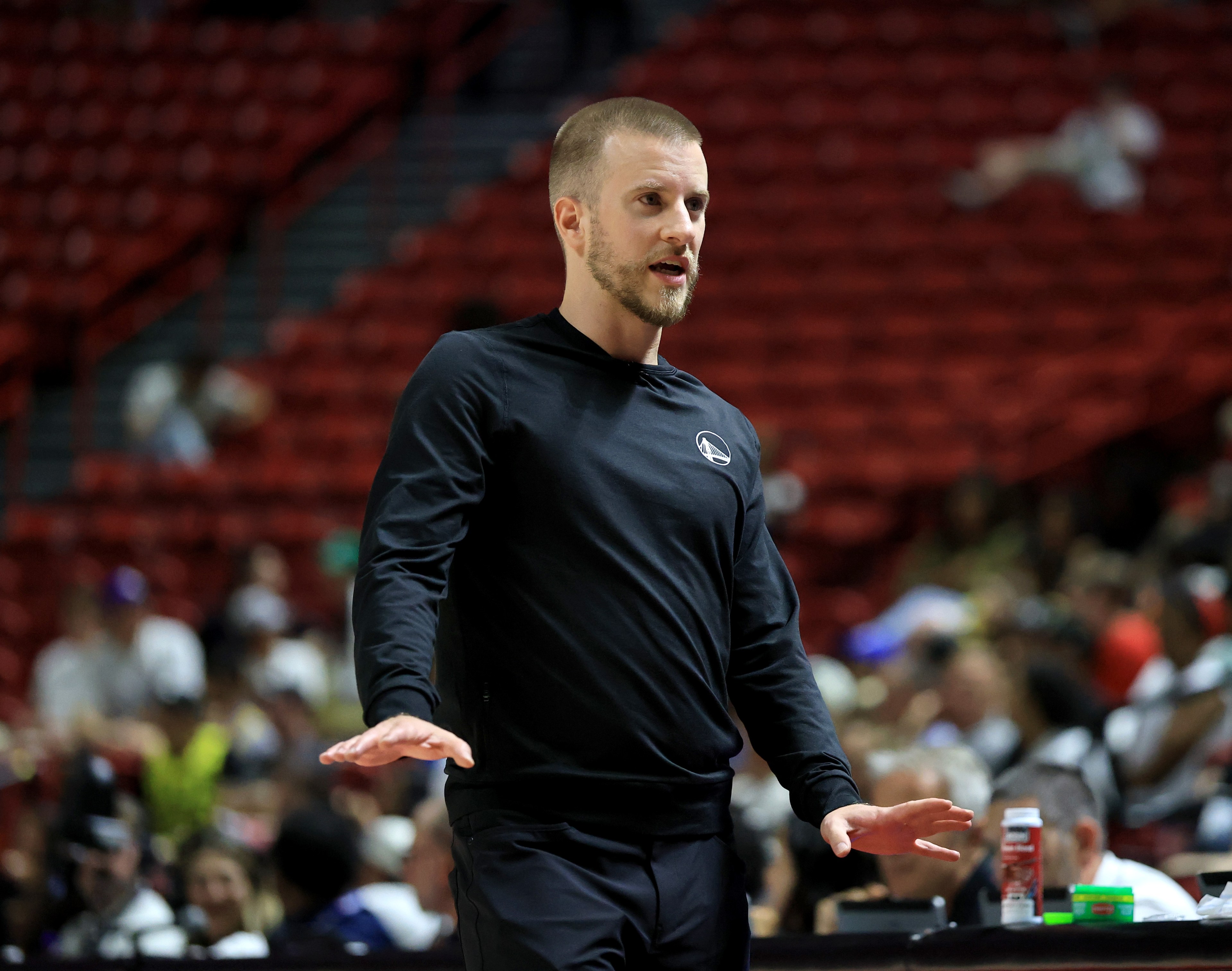 A man in a black long-sleeve shirt is speaking passionately in an indoor setting with blurred spectators in the background.