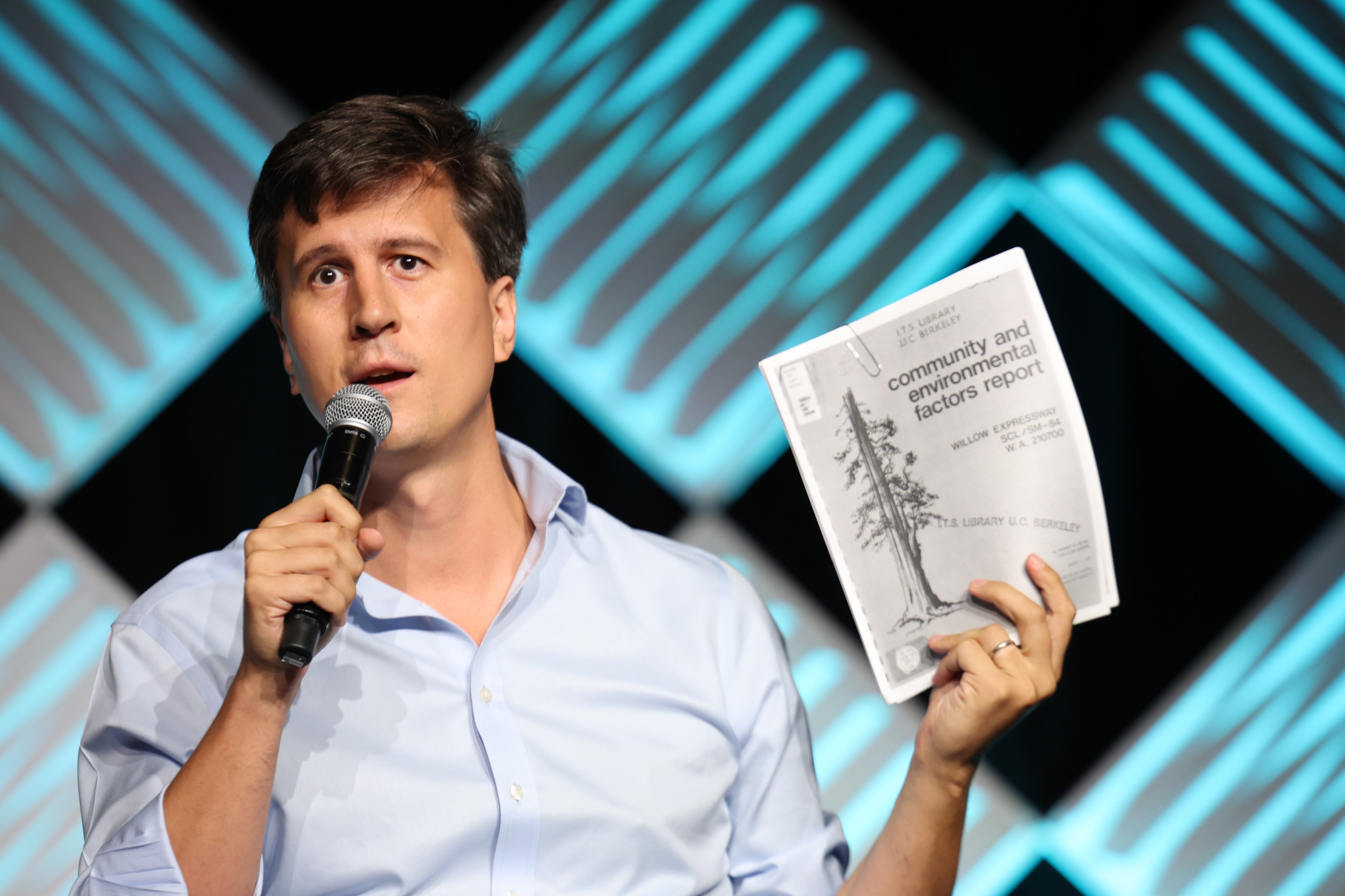 A man in a light blue shirt holds a microphone and a report titled &quot;Community and Environmental Factors Report.&quot; The background features a geometric pattern.