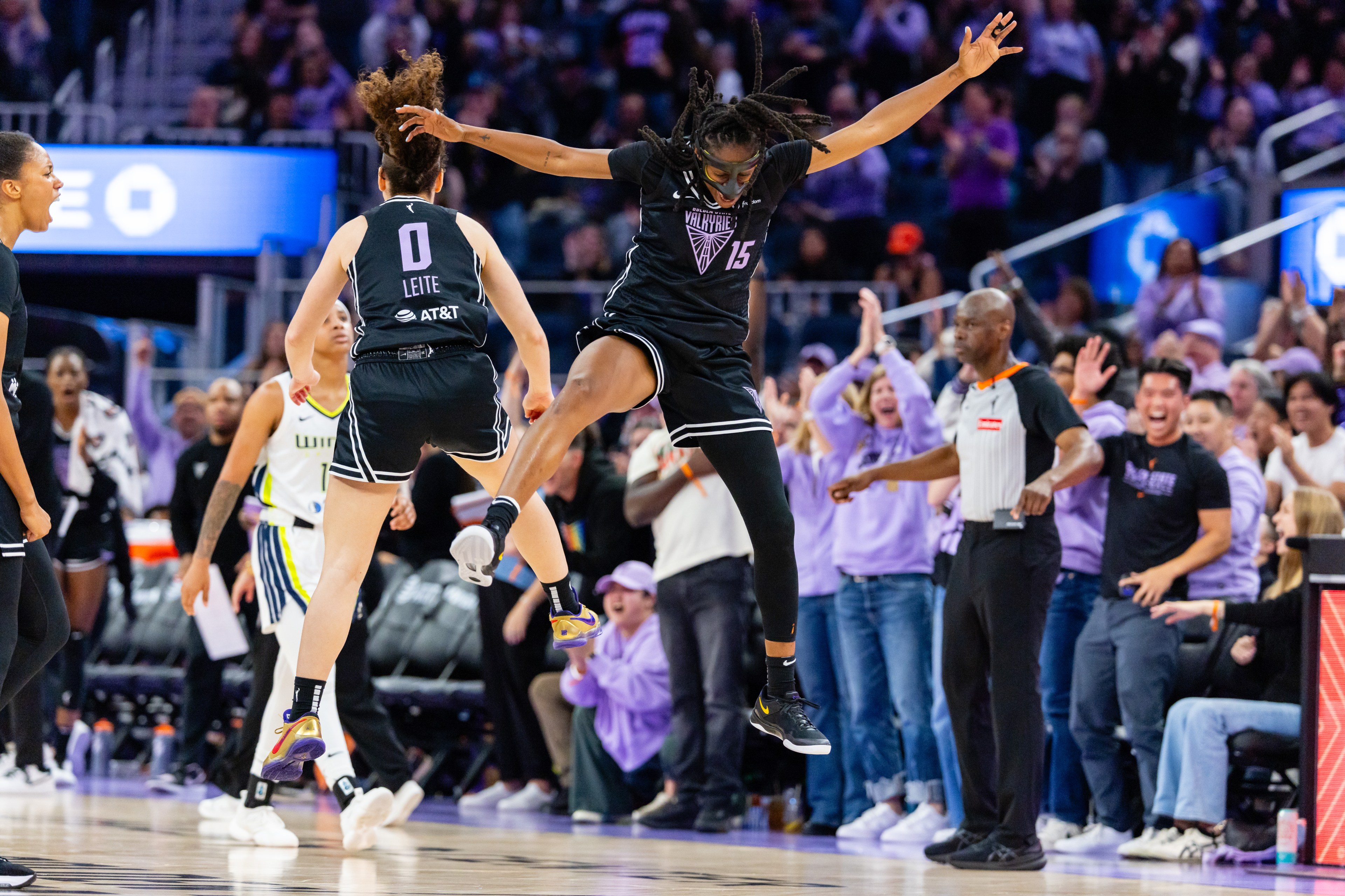 A basketball player in black jumps high, celebrating with teammates. The crowd in the background is cheering, creating an energetic atmosphere.