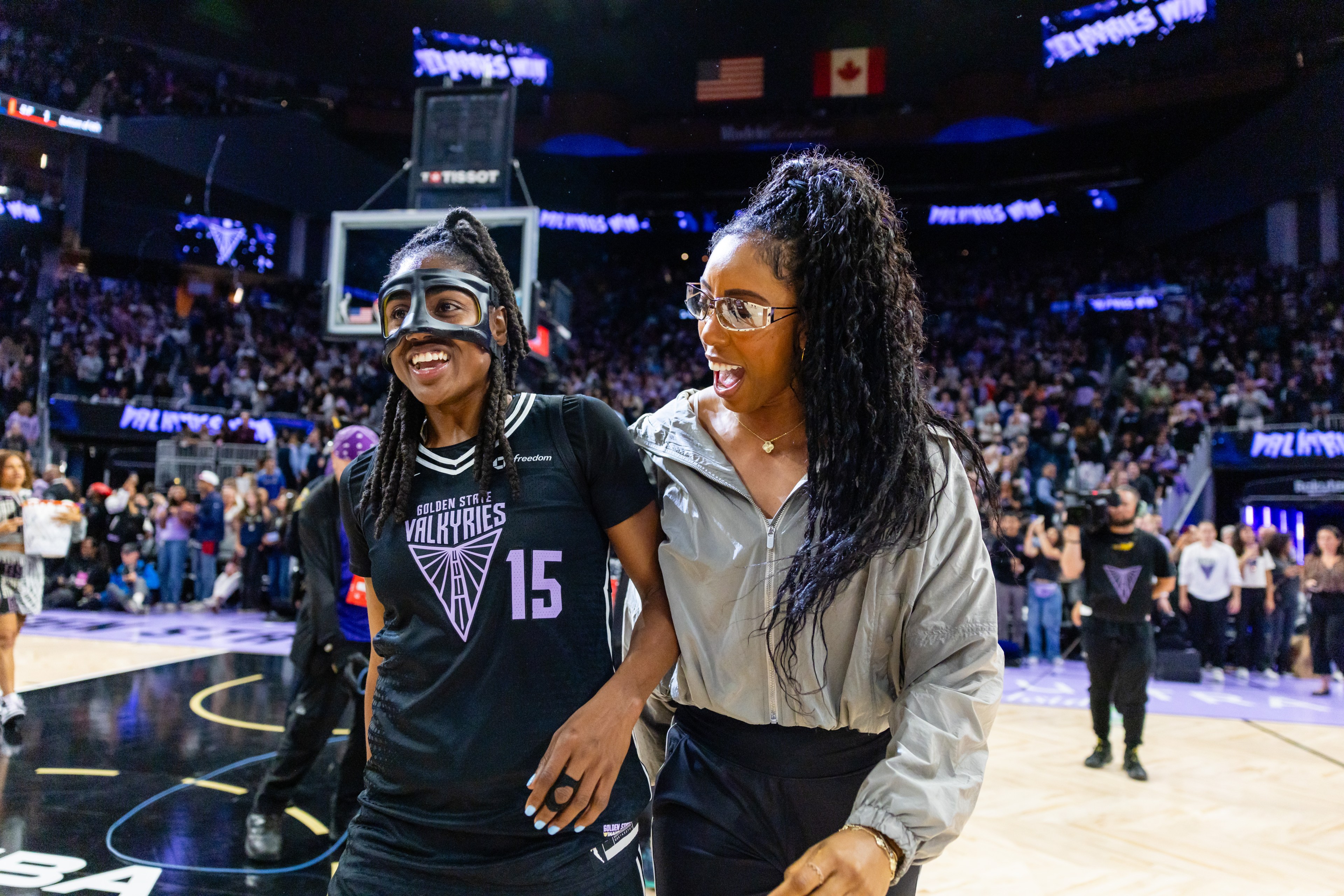 Two women on a basketball court are smiling. One wears a jersey and mask with "Valkyries" on it, the other a silver jacket. The crowd is cheering around them.