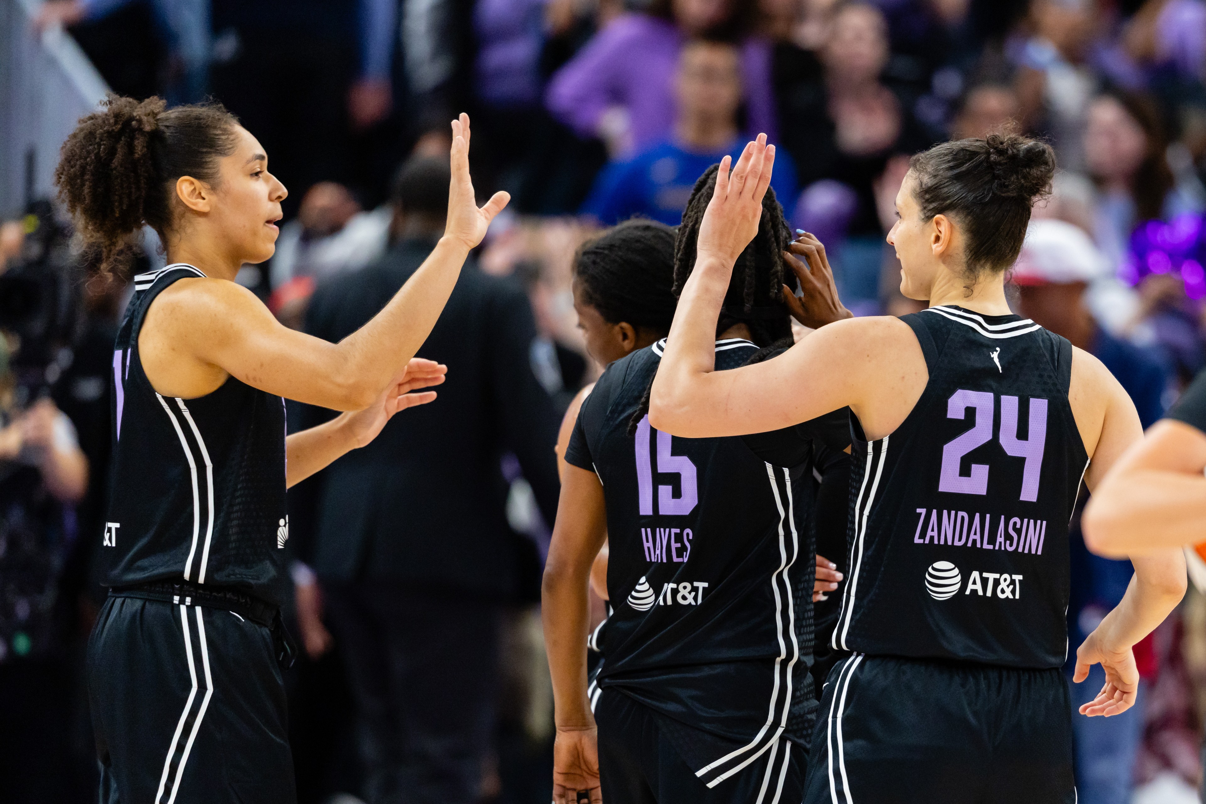 The image shows basketball players in black jerseys giving each other high-fives on the court. The focus is on teamwork and camaraderie during a game.