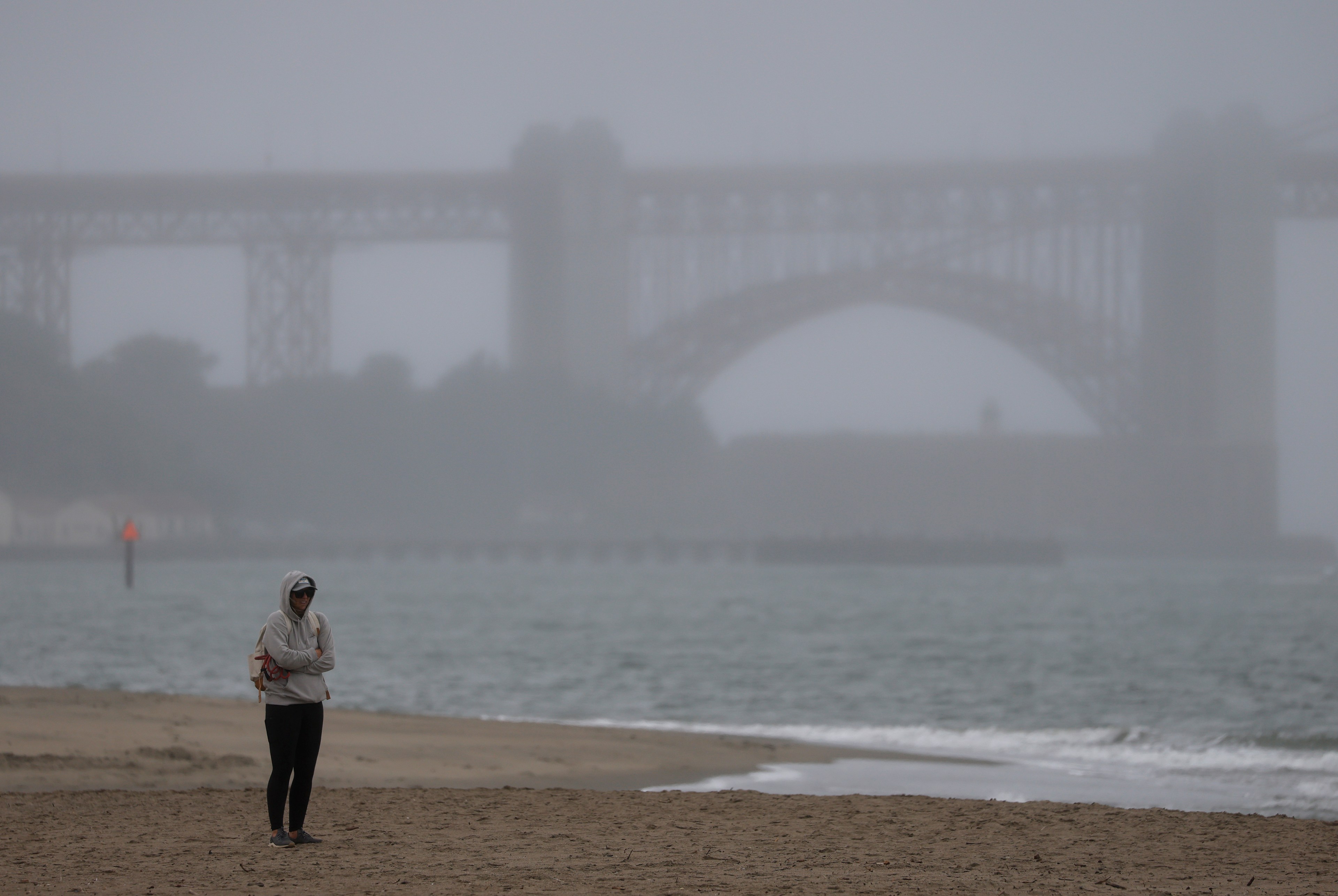 A person in a hoodie walks on a foggy beach near the shoreline. In the background, a large bridge looms faintly in the mist.