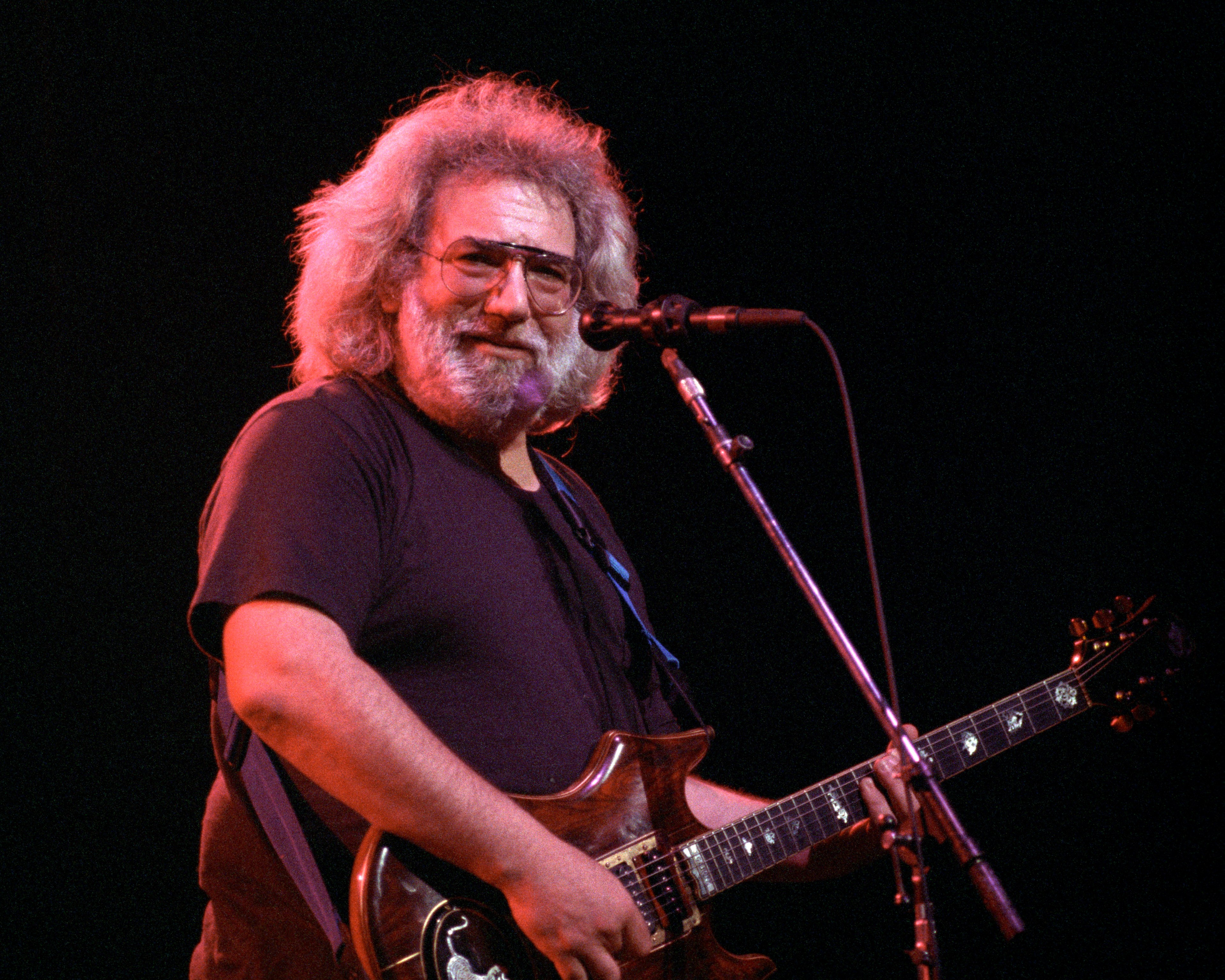 A bearded man with glasses plays an electric guitar on stage, smiling towards the audience. The background is dark, highlighting him under warm lighting.