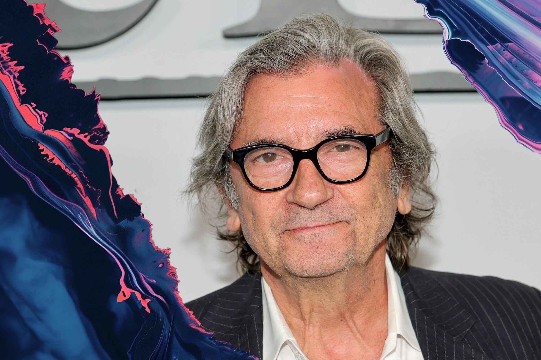 An older person with gray hair and glasses is smiling subtly. They are wearing a white shirt under a pinstripe jacket. The left side has a colorful, abstract effect.