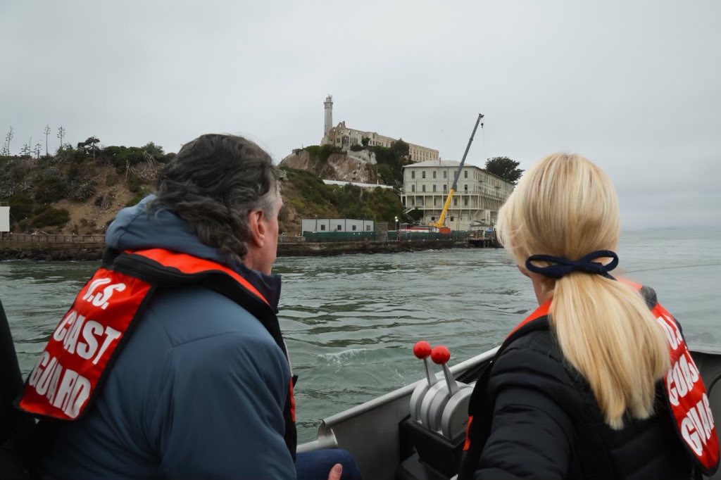 Two people in U.S. Coast Guard gear are on a boat near an island with a large, historic building and a crane. The sky is overcast.