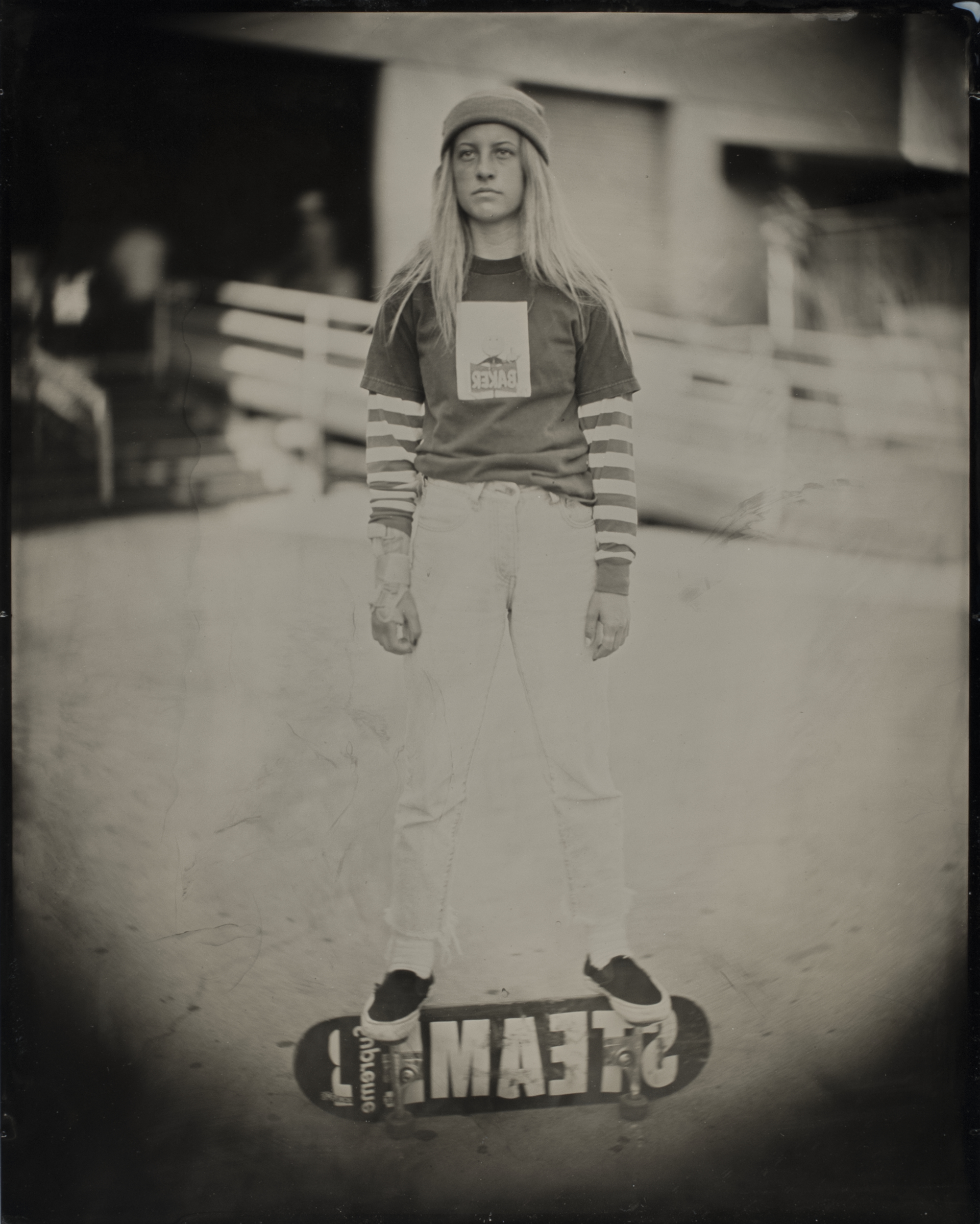 A person stands on a skateboard labeled &quot;TEAM&quot; wearing a beanie, striped long sleeves under a t-shirt, and jeans. The background is blurred and urban.