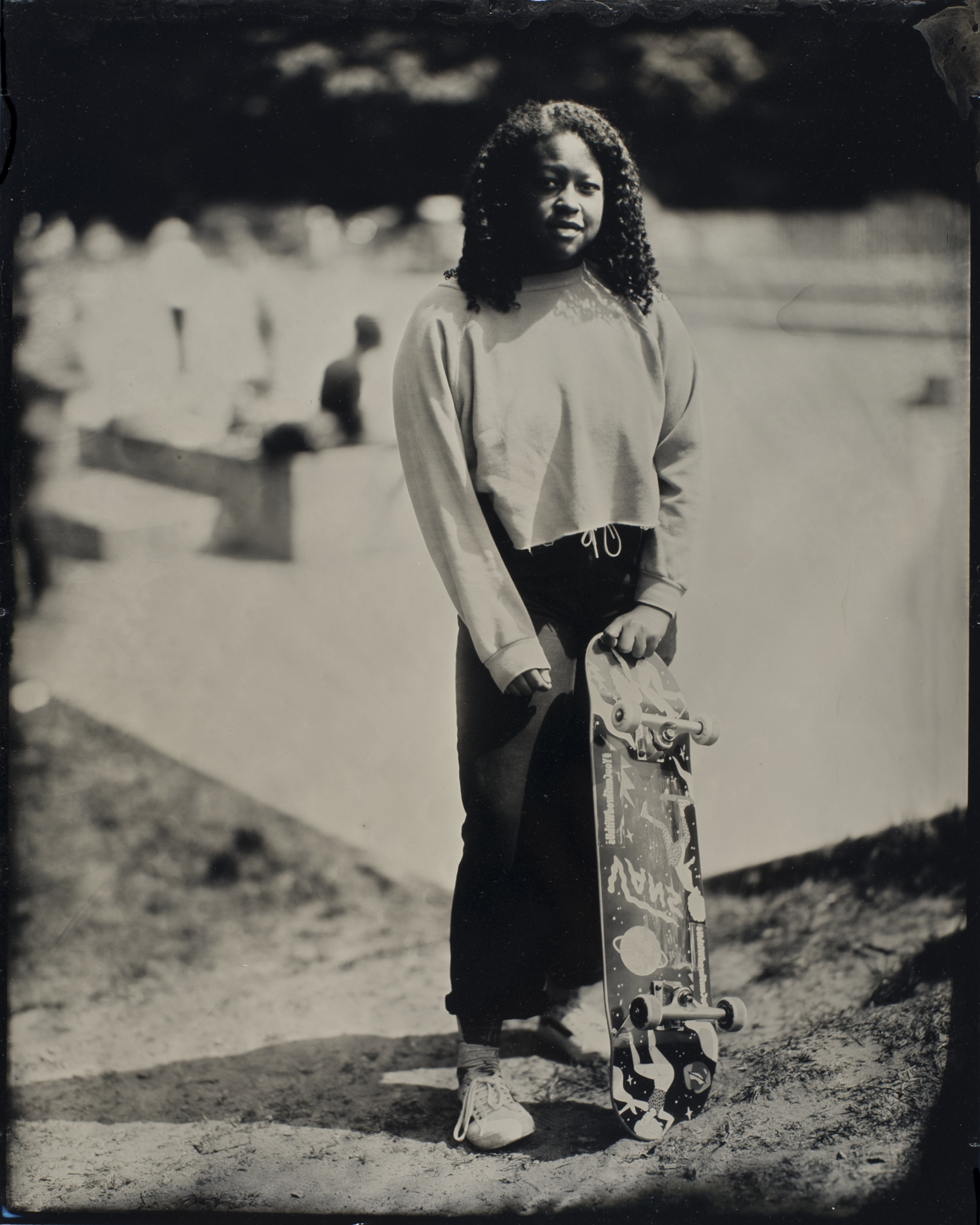 A person stands outdoors holding a skateboard vertically. They're wearing a light sweater and pants, with a blurred background of a skatepark and trees.