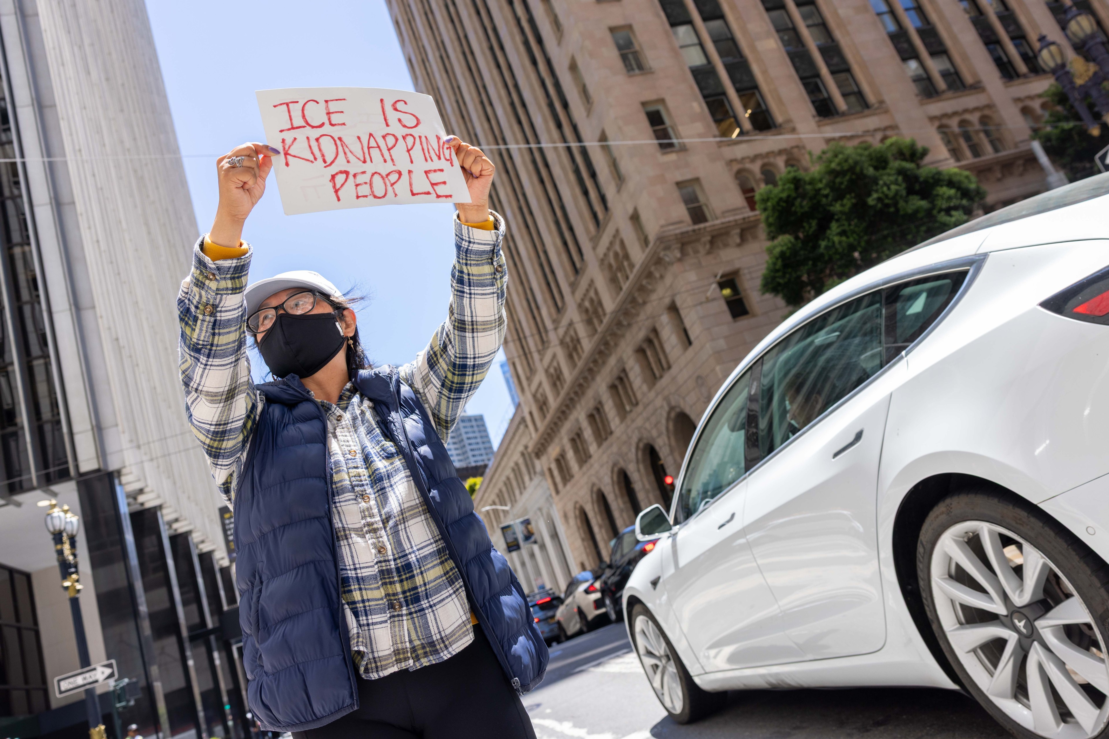 A person wearing a mask holds a sign saying "ICE IS KIDNAPPING PEOPLE" on a city street, with tall buildings and a white car in the background.