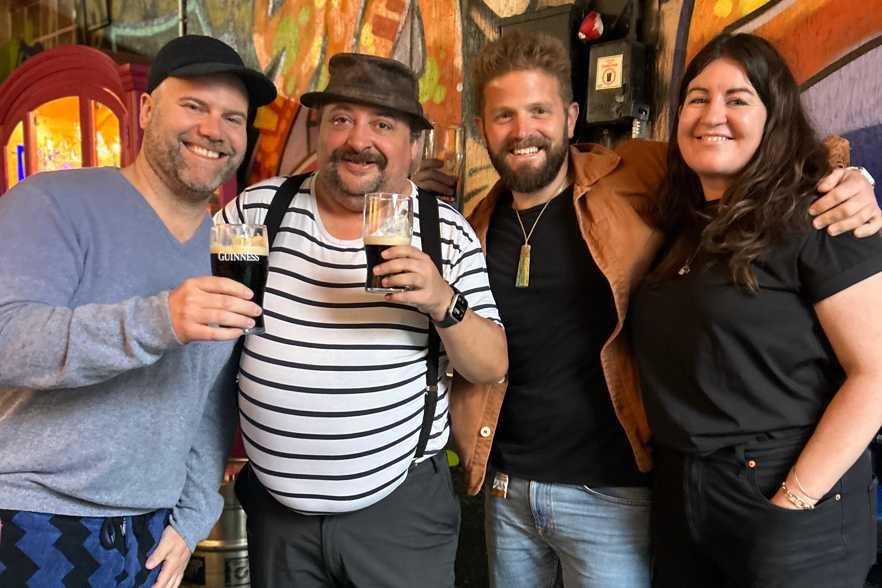 Four people stand closely together, smiling. Two hold dark beer glasses. They're in front of a colorful graffiti mural with vibrant, abstract designs.